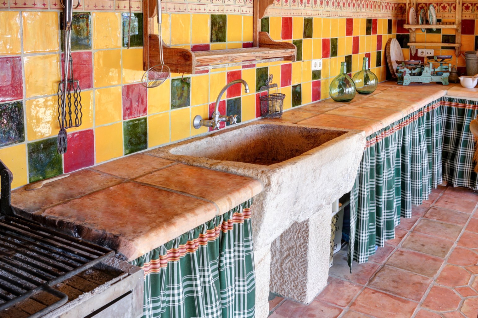 Cuisine d'été avec évier en pierre massive, plan de travail en terre cuite et crédence en céramique colorée. Comprend un espace barbecue et des rideaux de rangement traditionnels.
