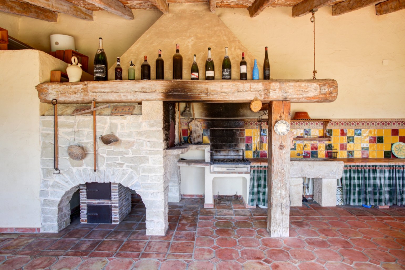 Cuisine d'été rustique avec barbecue intégré, imposante poutre en bois ancien, évier en pierre naturelle et crédence en faïence colorée sur sol en terre cuite.