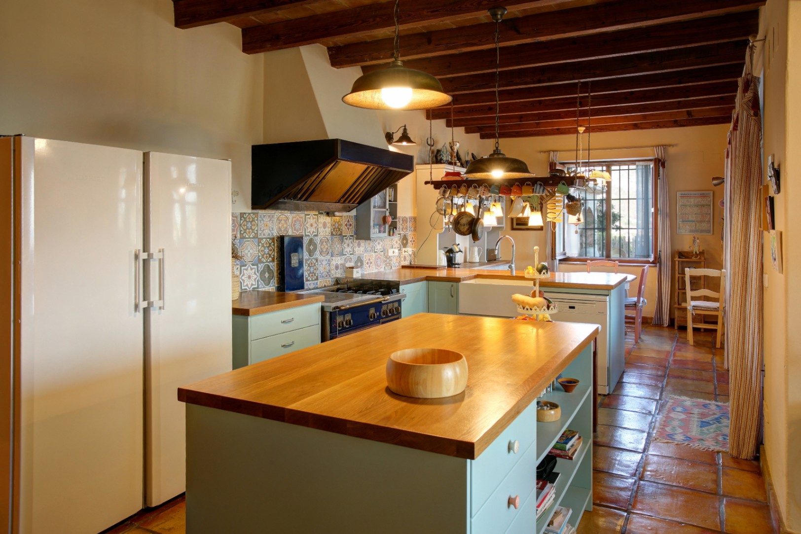 Cuisine rustique avec îlot central en bois, poutres apparentes et sol en terre cuite. Elle dispose d'une cuisinière professionnelle bleue et d'une crédence en carreaux décoratifs.
