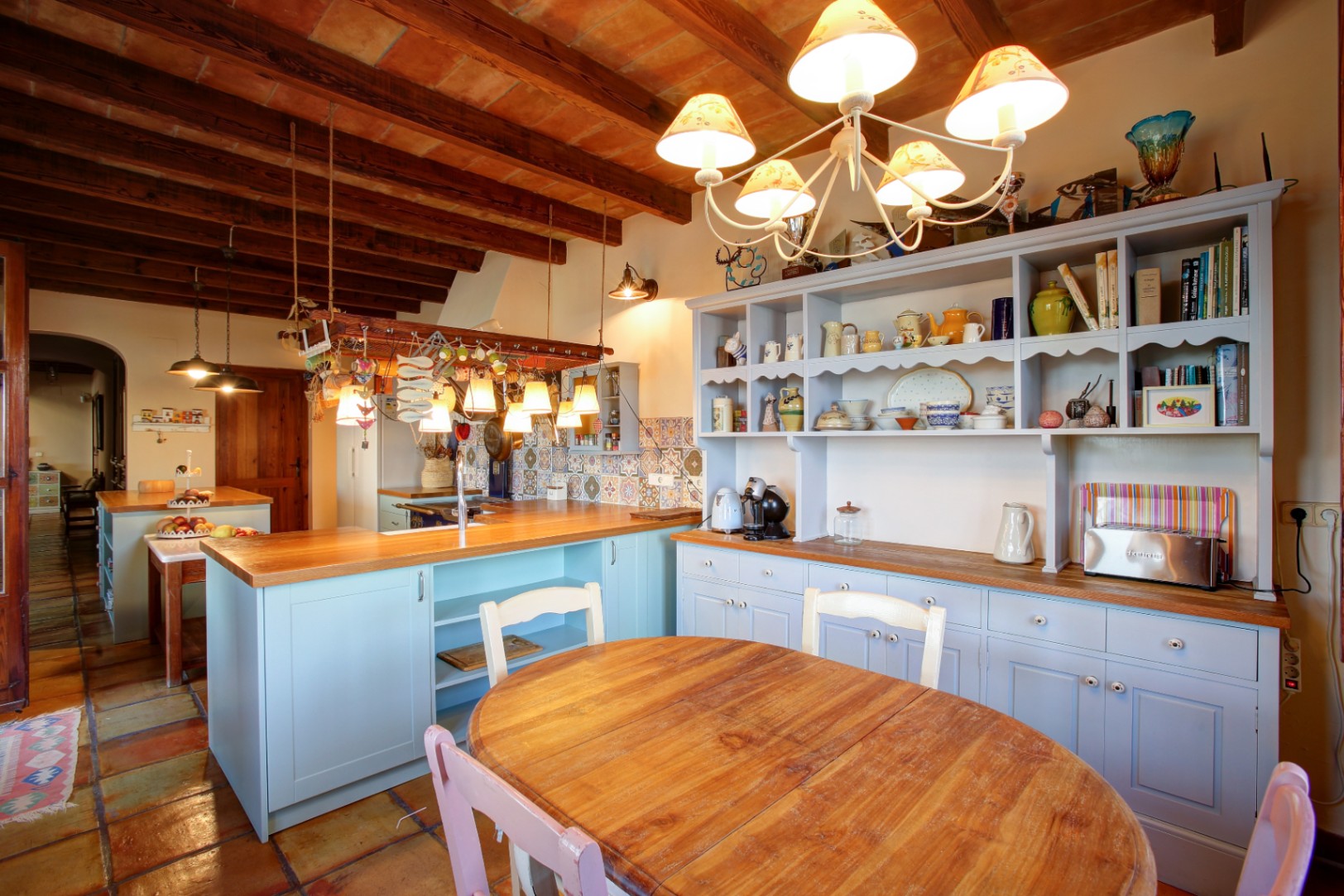 Cuisine rustique avec meubles bleu clair, plans de travail en bois et plafonds à poutres apparentes. Comprend un coin repas avec sol en terre cuite traditionnelle.