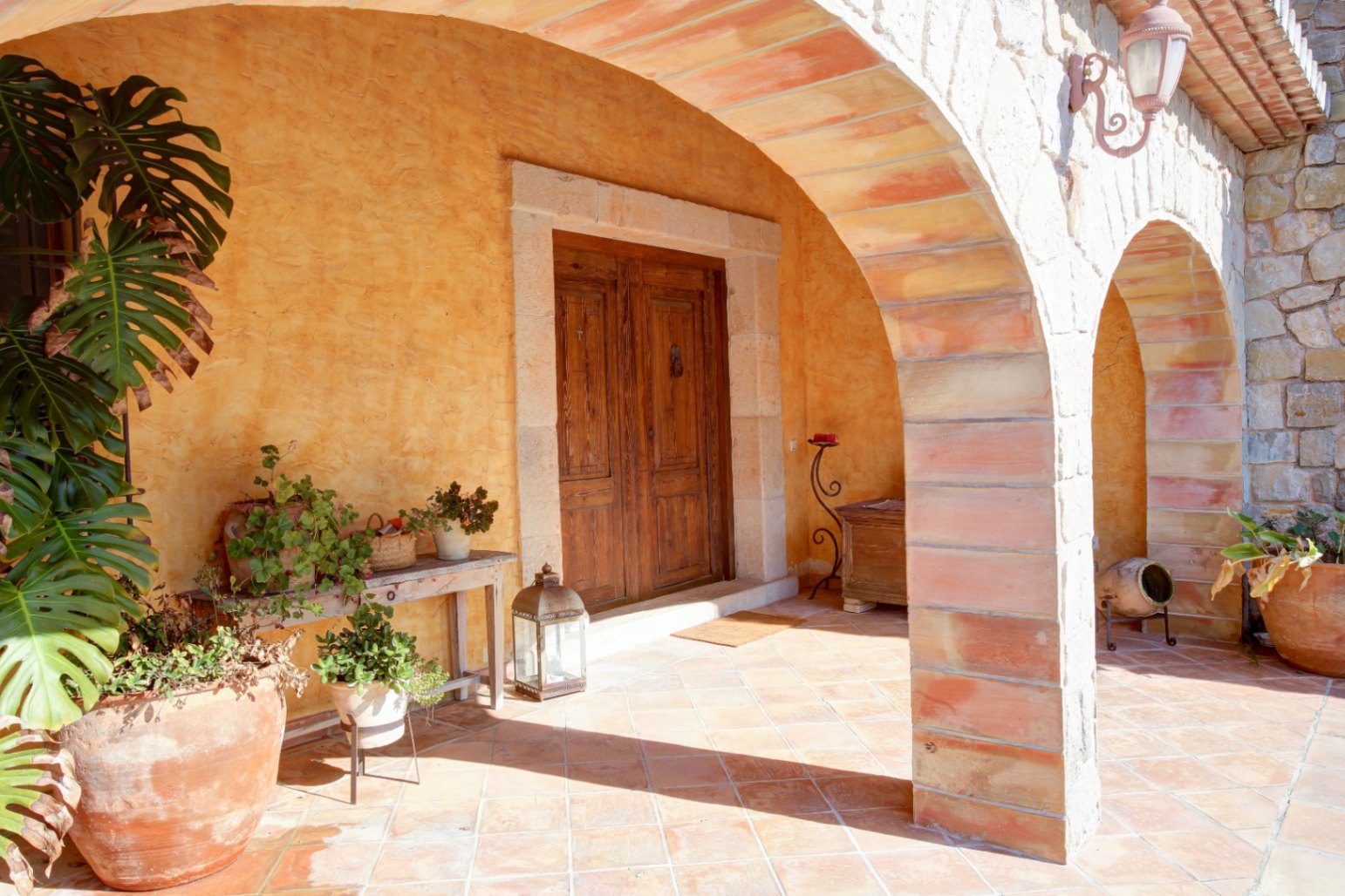Entrée principale sous un porche aux arches de pierre et brique, avec sol en terre cuite, porte en bois massif et plantes méditerranéennes décoratives.