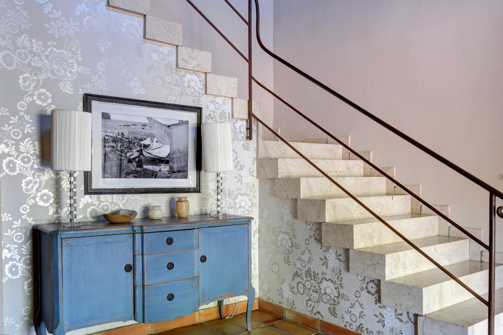 Escalier en marbre massif avec rampe métallique, accompagné d'un buffet bleu patiné, d'un papier peint floral et d'un sol en terre cuite.