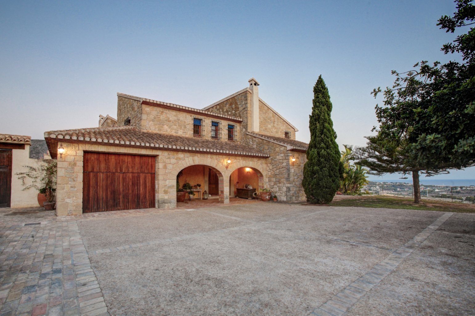Façade en pierre naturelle avec arches rustiques, porte de garage en bois massif et vaste cour pavée offrant une vue dégagée sur la mer Méditerranée.