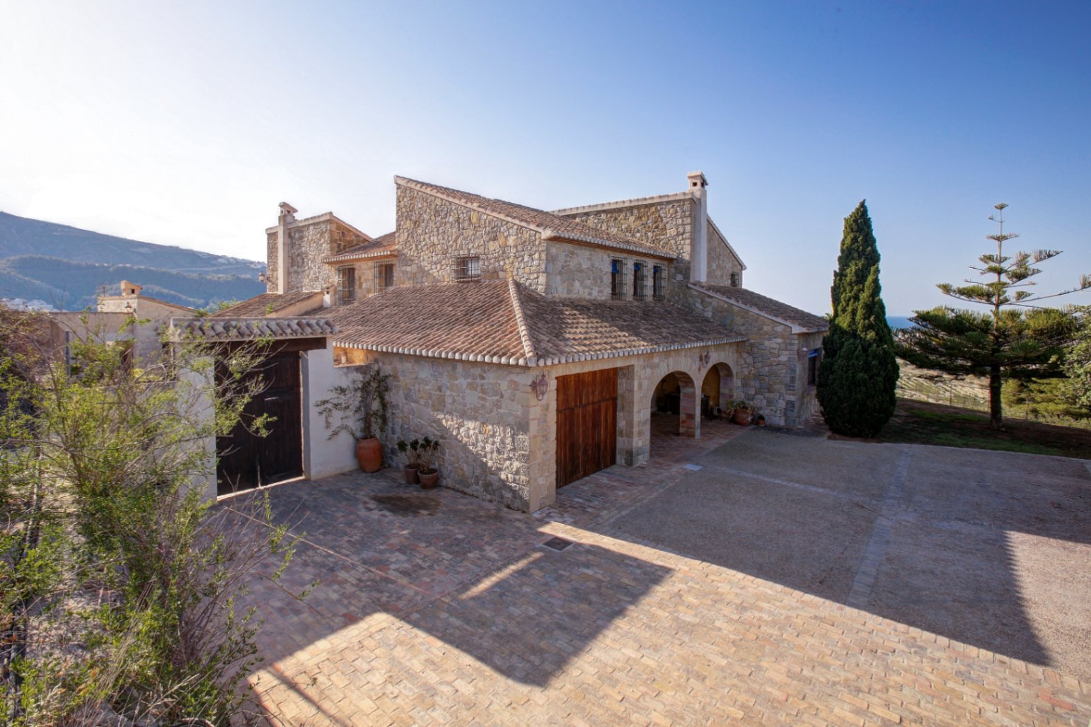 Façade en pierre naturelle avec arches traditionnelles, toiture en tuiles et vaste cour pavée de terre cuite sous le soleil méditerranéen.