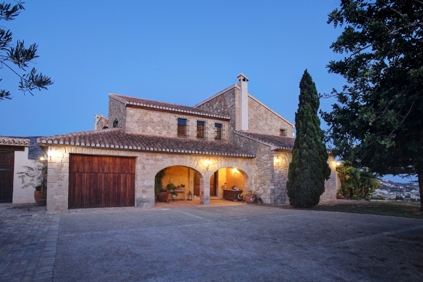 Façade en pierre naturelle avec porches en arche et porte de garage en bois, mise en valeur par un éclairage crépusculaire sur une cour pavée.