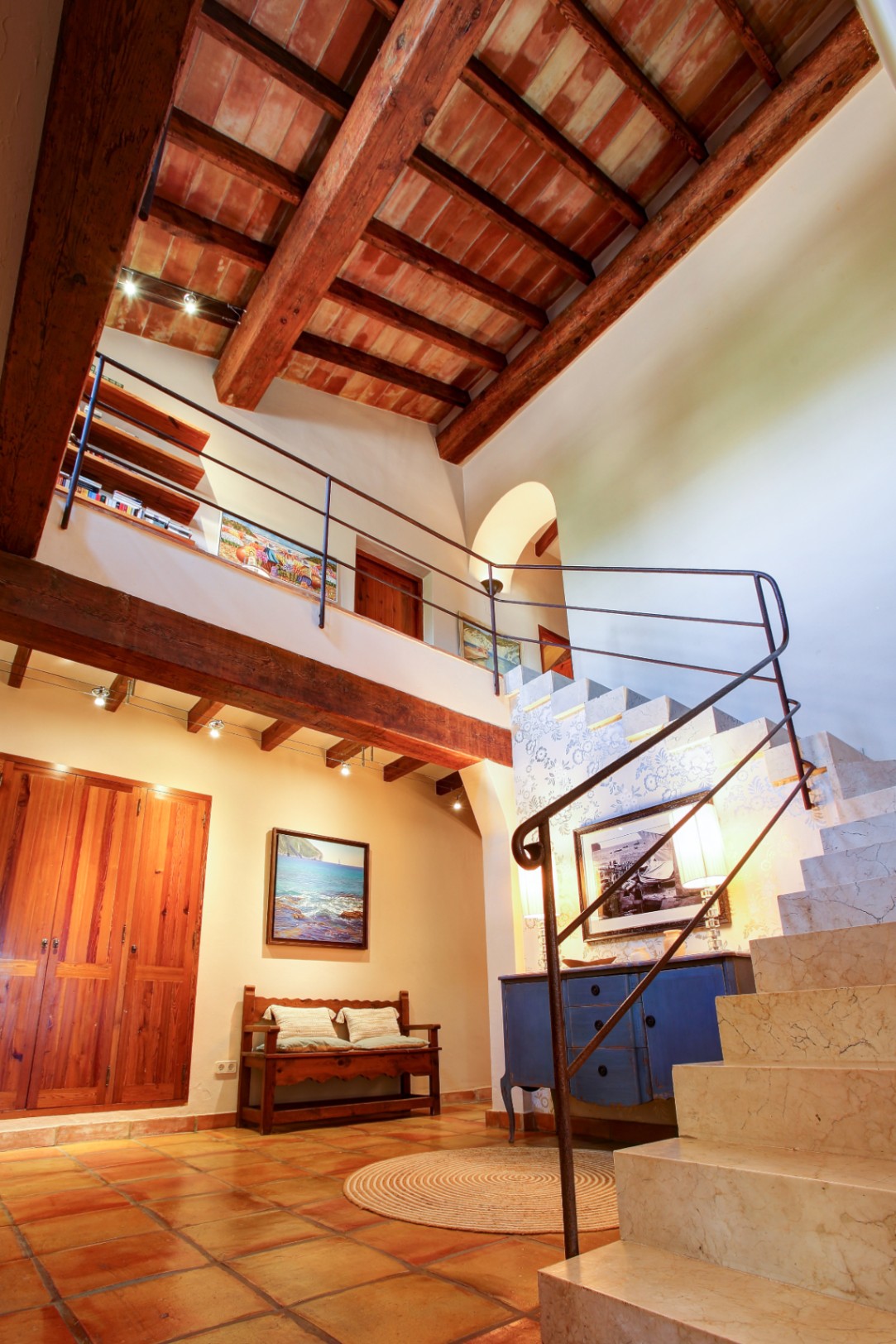 Hall d'entrée avec plafonds à double hauteur, poutres en bois apparentes et terre cuite. L'escalier en marbre massif et le sol en terre cuite soulignent le style rustique.