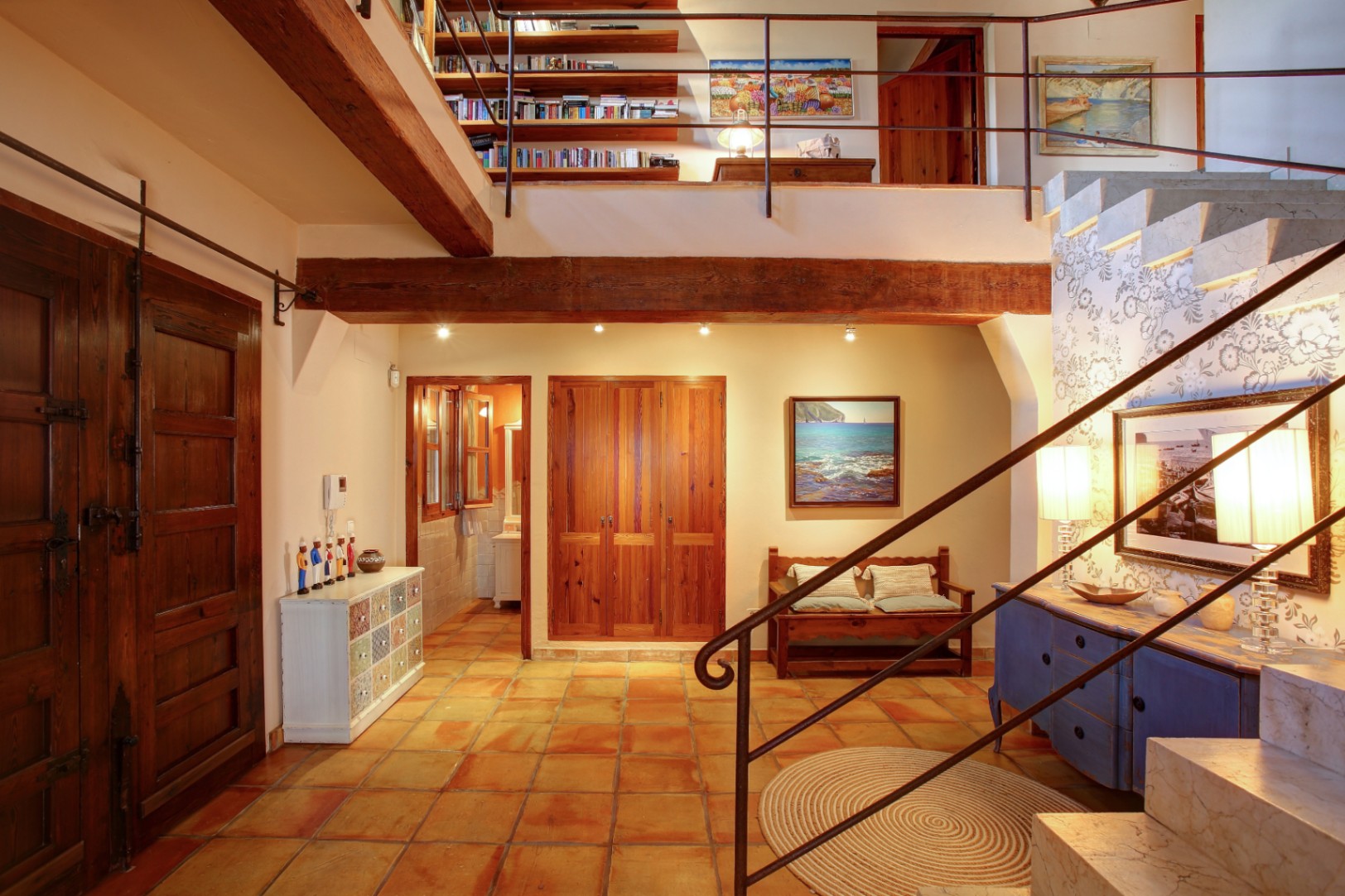 Hall d'entrée avec sols en terre cuite, poutres en bois massif et un escalier en marbre menant à une mezzanine aménagée en bibliothèque.