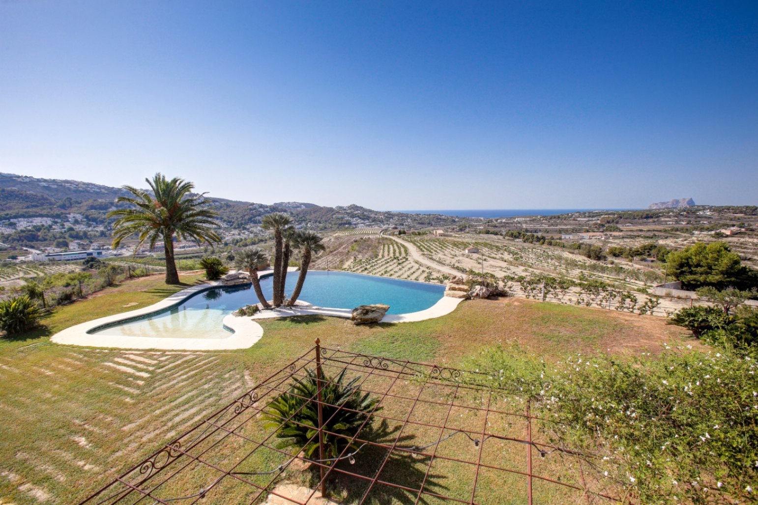 Piscine à débordement aux formes organiques offrant une vue dégagée sur la mer et le Peñón de Ifach, entourée de jardins et de vignobles.