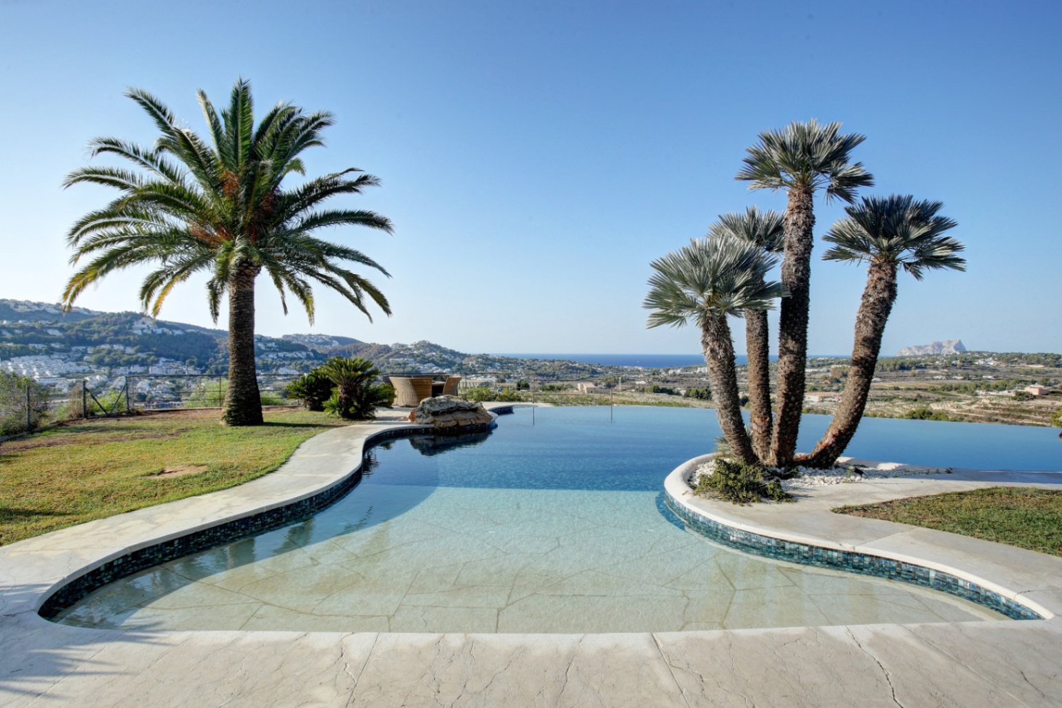 Piscine à débordement de forme organique bordée de palmiers, offrant une vue panoramique sur la mer Méditerranée et les collines environnantes de Moraira.