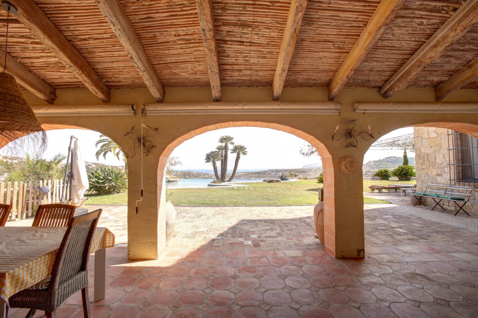 Porche couvert avec poutres en bois, plafond en canisse et sol en terre cuite. Les arches encadrent une vue sur le jardin, la piscine aux palmiers et la mer.