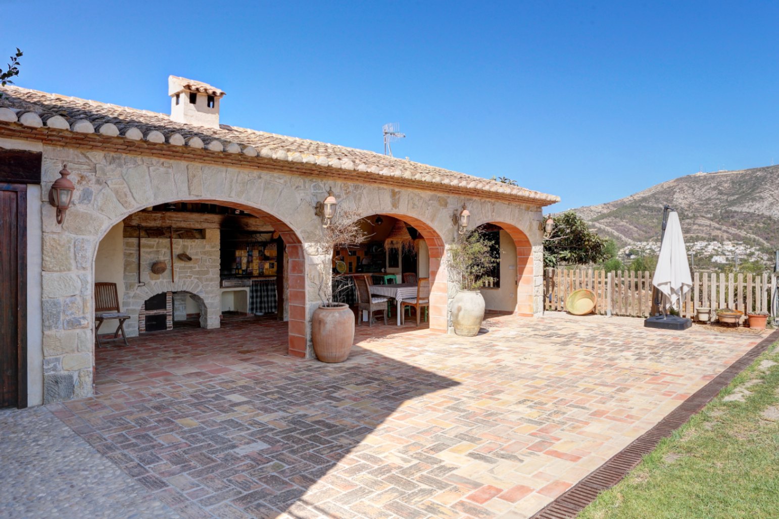 Porche rustique avec arches en pierre, sol en terre cuite et espace barbecue traditionnel intégré offrant une vue dégagée sur les montagnes.