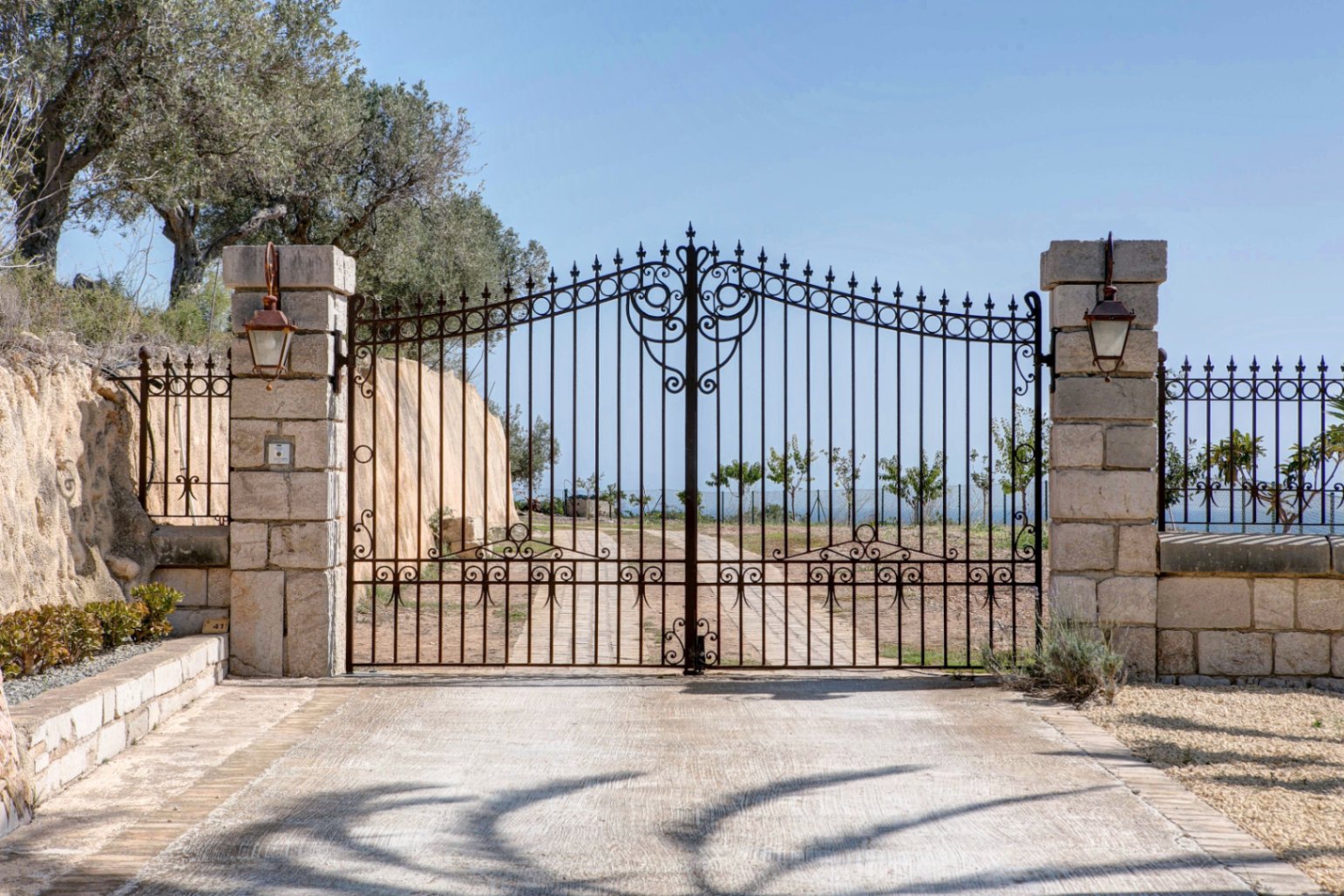 Portail d'entrée en fer forgé travaillé, encadré par des piliers en pierre naturelle avec lanternes classiques et une allée pavée privée.