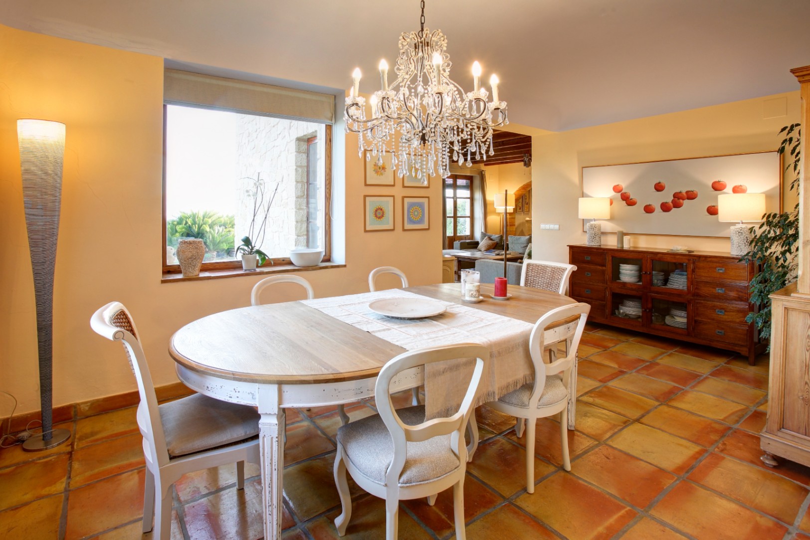 Salle à manger avec sol en terre cuite, table en bois décapé et lustre en cristal. La fenêtre illumine l'espace et offre une vue sur les murs en pierre extérieurs.