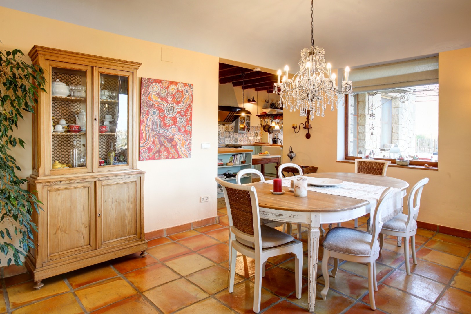 Salle à manger avec table en bois, chaises patinées et lustre en cristal, complétée par un sol en terre cuite et un buffet en bois naturel.