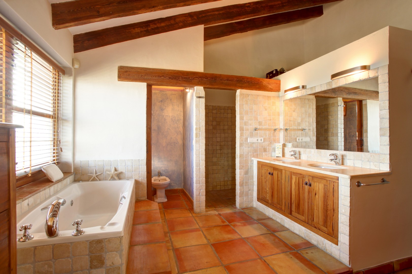 Salle de bain rustique avec poutres en bois apparentes, sol en terre cuite et baignoire encastrée. Le meuble double vasque allie bois de mobila, plan de travail en marbre et finitions en pierre.