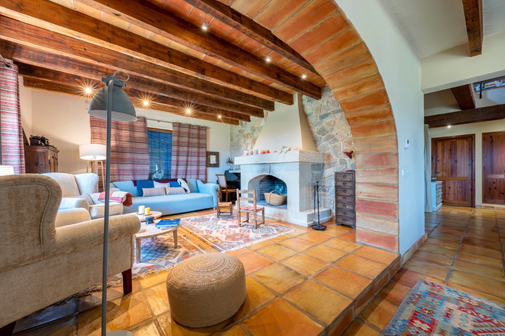 Salon rustique avec cheminée en pierre, plafond aux poutres apparentes et terre cuite, sols en terre cuite et une grande arche décorative en briques.