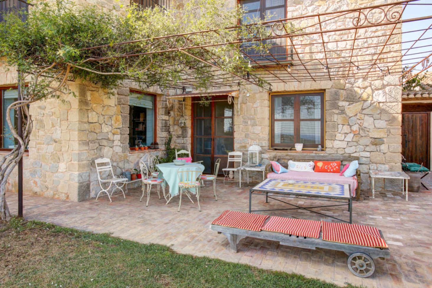 Terrasse extérieure avec façade en pierre naturelle, sol en terre cuite et pergola en fer forgé. L'espace offre un coin salon rustique et une table basse avec plateau en mosaïque.