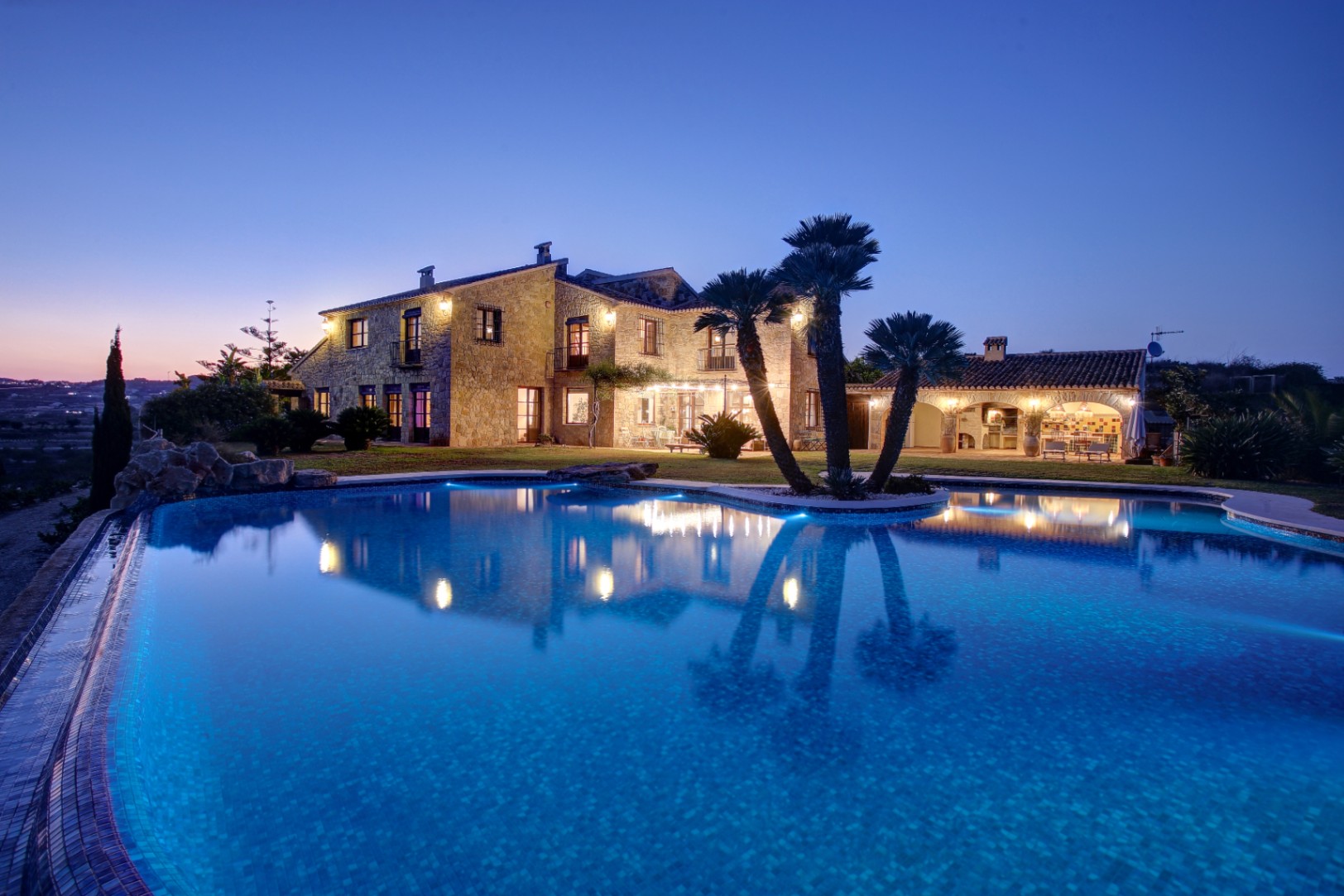 Vue au crépuscule de la façade en pierre et de la piscine éclairée, incluant la cuisine d'été et des palmiers intégrés dans l'aménagement paysager.