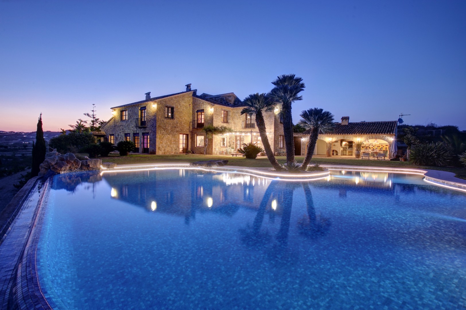 Vue nocturne de la façade en pierre naturelle et de la piscine organique avec éclairage LED, incluant la cuisine d'été et l'espace barbecue extérieur.