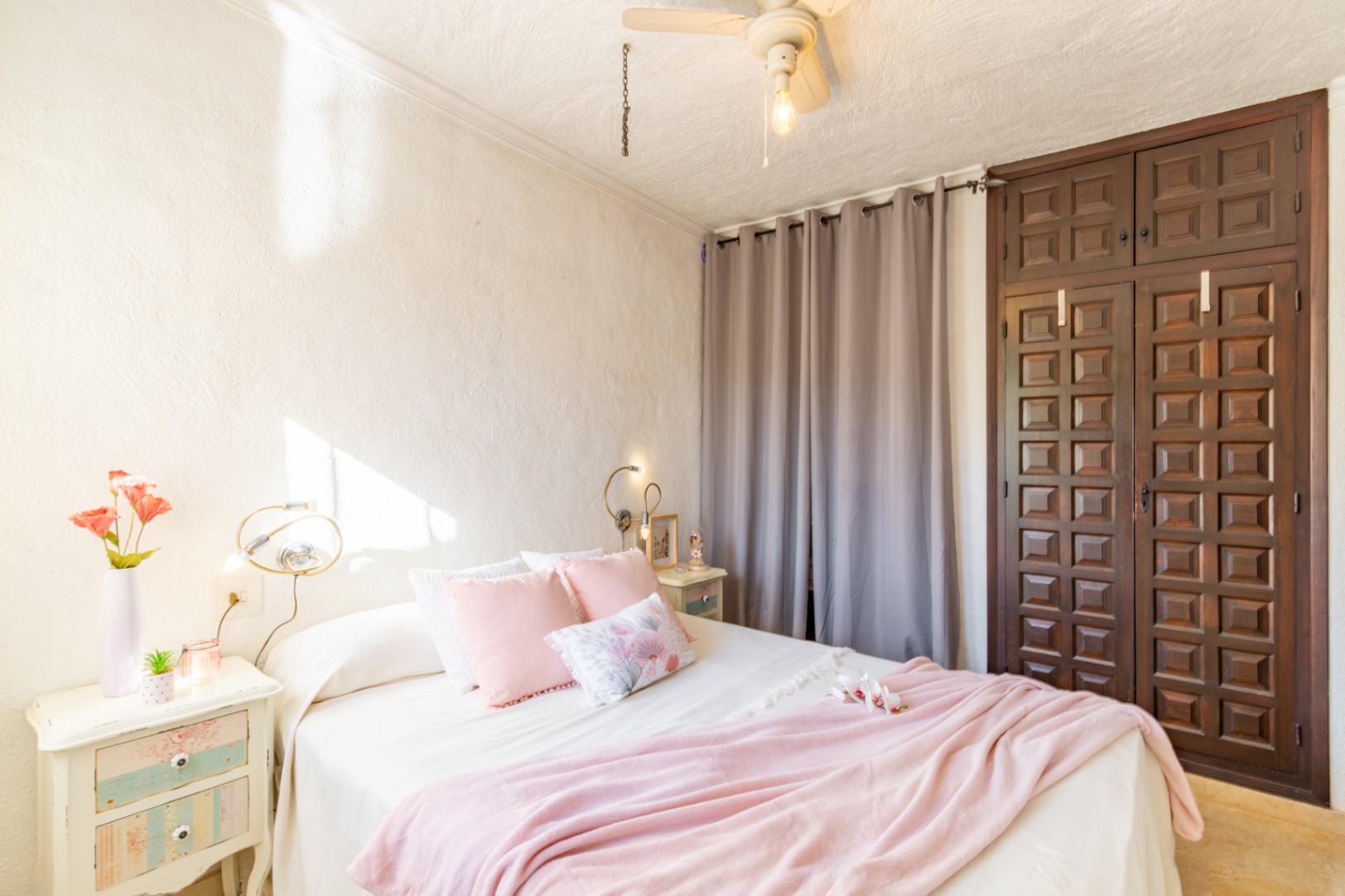 Chambre avec armoire encastrée en bois sombre sculpté, murs texturés blancs et ventilateur de plafond. La pièce bénéficie d'une belle luminosité naturelle.