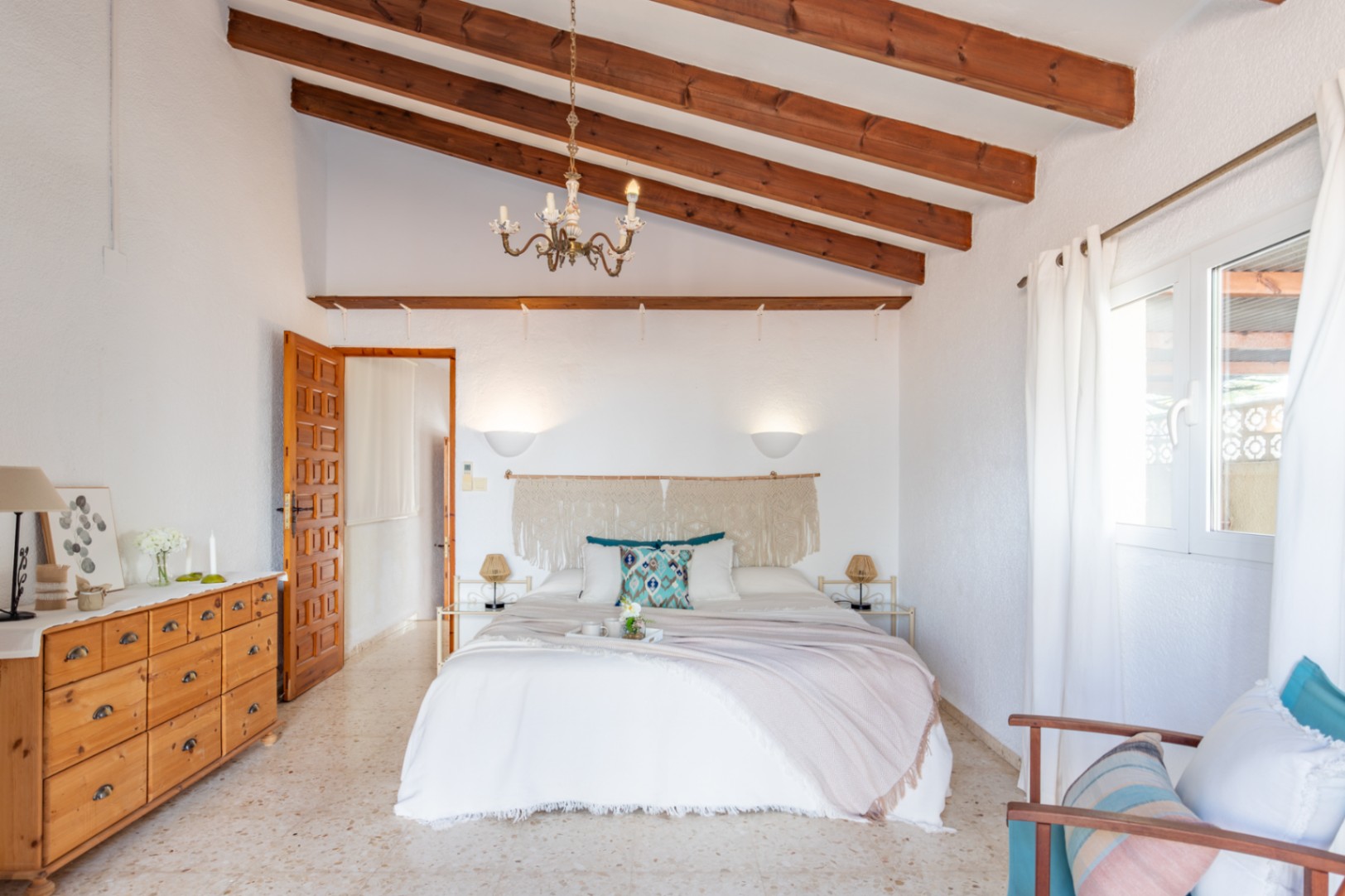 Chambre avec hauts plafonds aux poutres en bois apparentes, sol en terrazzo et décoration méditerranéenne avec tête de lit en macramé et mobilier en bois.