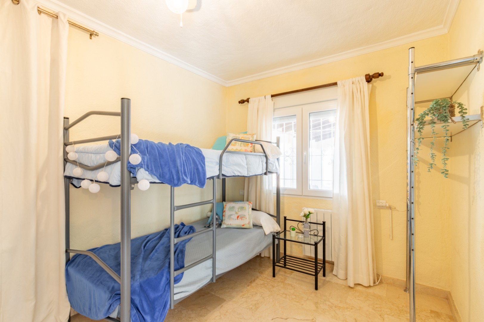 Chambre avec lits superposés en métal, murs crème et sol en marbre poli. La pièce dispose d'une fenêtre lumineuse et d'un système de rangement ouvert.