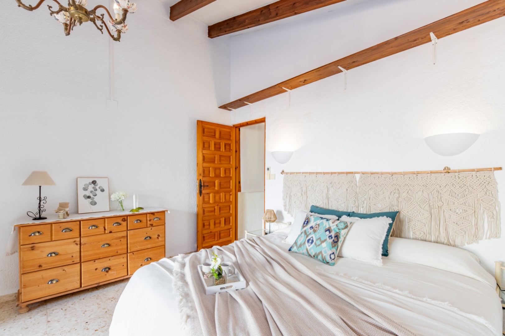 Chambre avec poutres en bois apparentes, commode en pin et tête de lit en macramé. Elle dispose d'un sol en terrazzo d'origine et d'une porte en bois sculpté de style rustique.