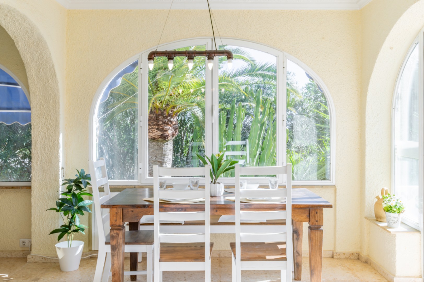 Coin repas lumineux sous une terrasse vitrée avec table en bois, chaises blanches et luminaire industriel, offrant une vue sur le jardin tropical.