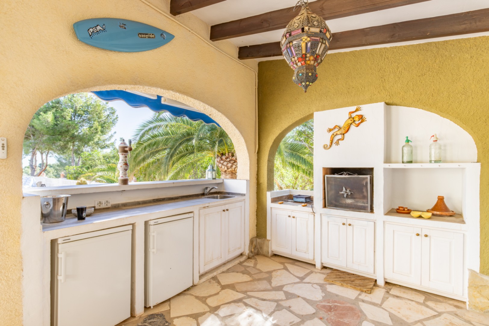 Cuisine d'été couverte avec poutres en bois, barbecue intégré, plusieurs réfrigérateurs et évier. Elle dispose d'un sol en pierre naturelle et d'une vue sur le jardin tropical.