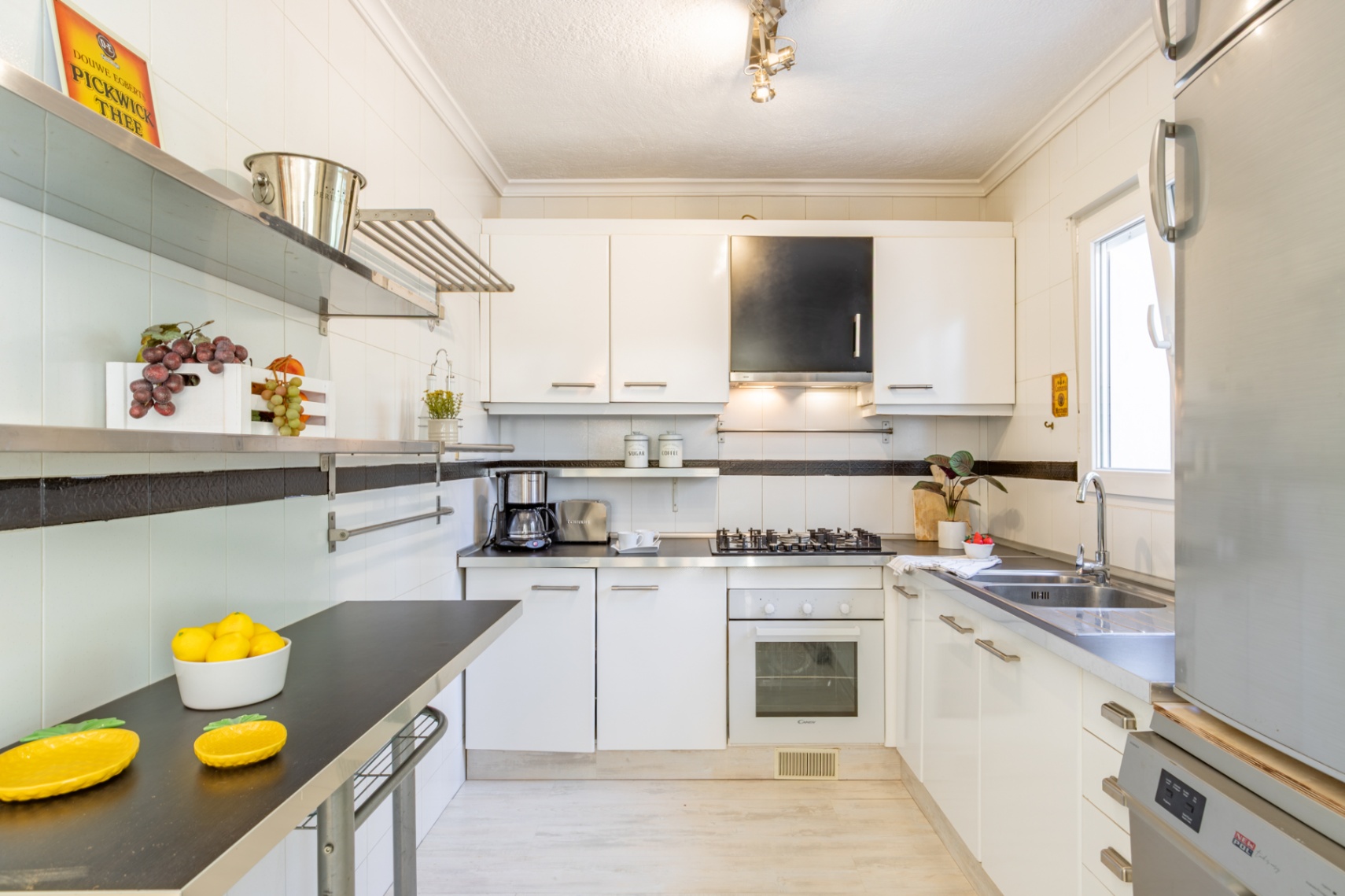 Cuisine équipée avec meubles blancs, plans de travail sombres, étagères en inox et lumière naturelle. Comprend une plaque de cuisson gaz, un four et un évier double.