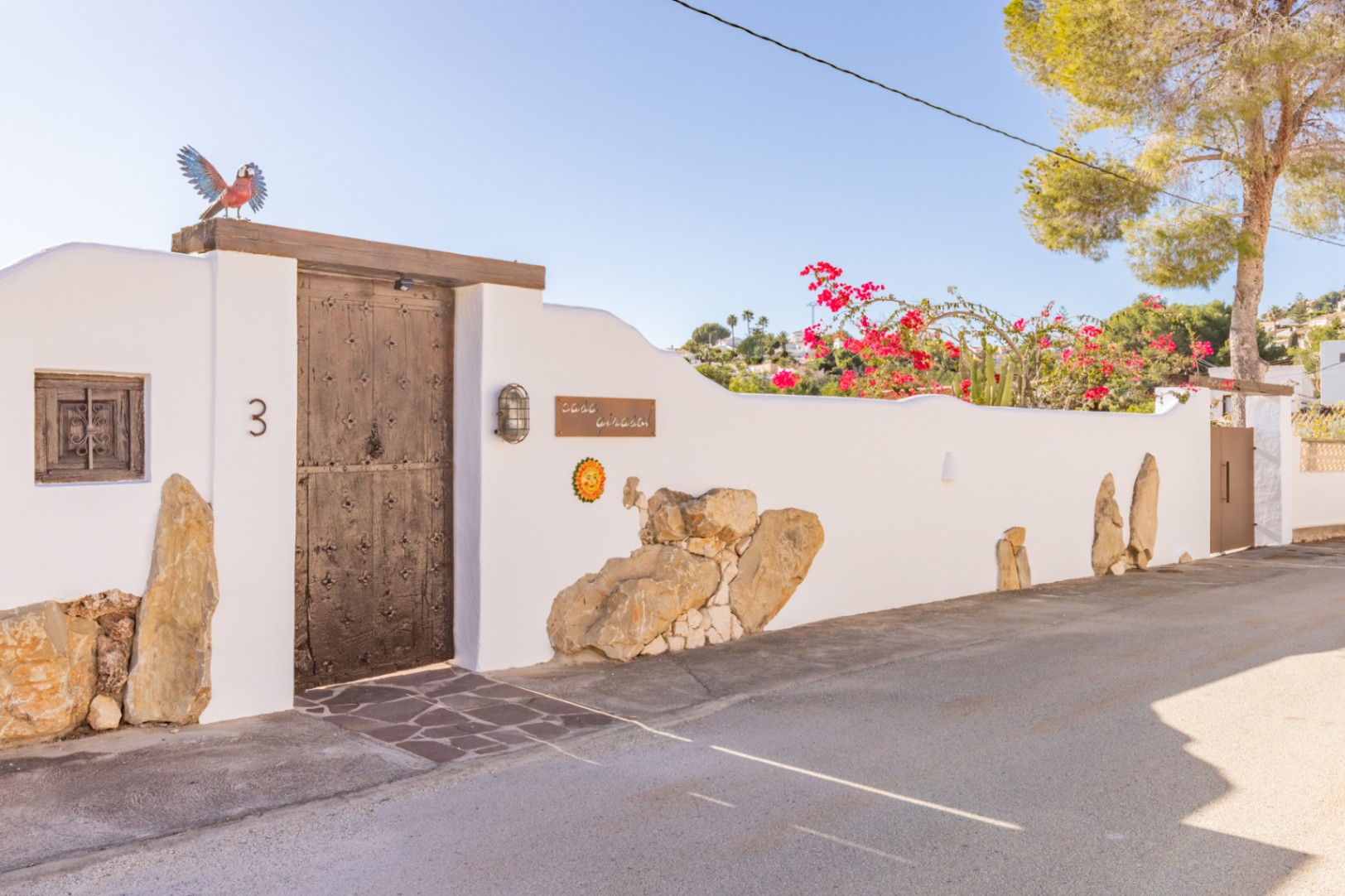 Entrée principale avec porte en bois rustique et ferrures, intégrée dans un mur blanc orné de pierres naturelles et de bougainvilliers en fleurs.