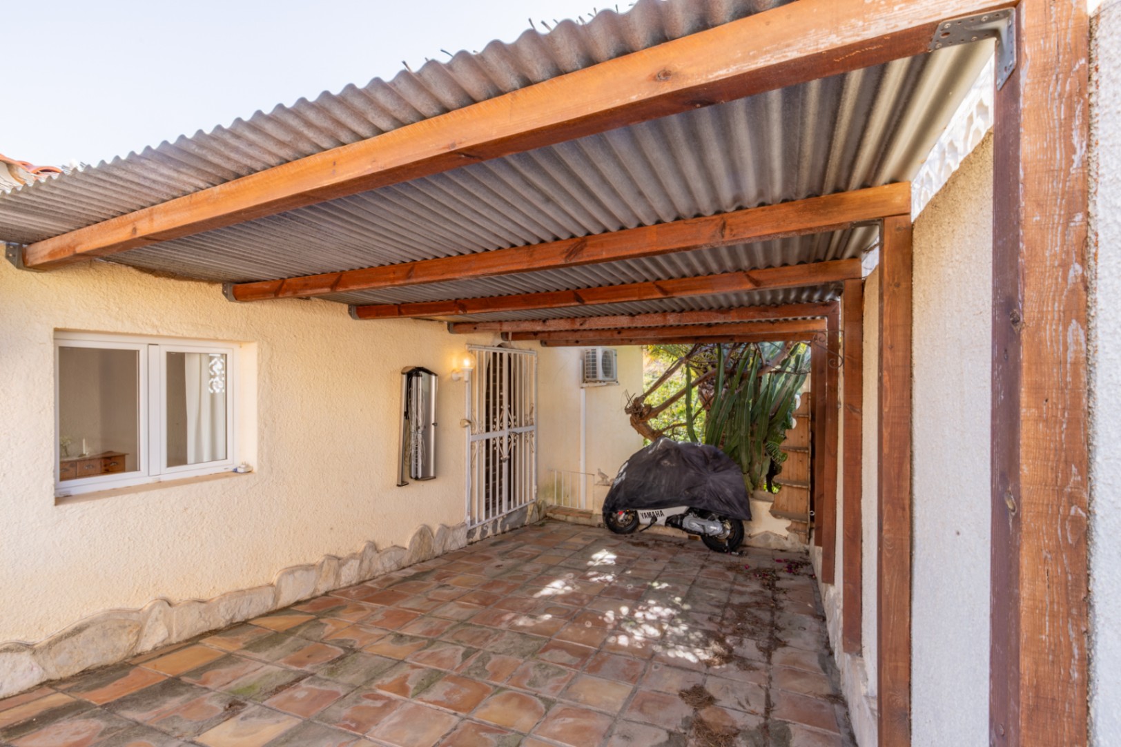 Espace de stationnement couvert avec charpente en bois, toiture ondulée et sol en grès rustique, offrant un accès direct et sécurisé à la propriété.