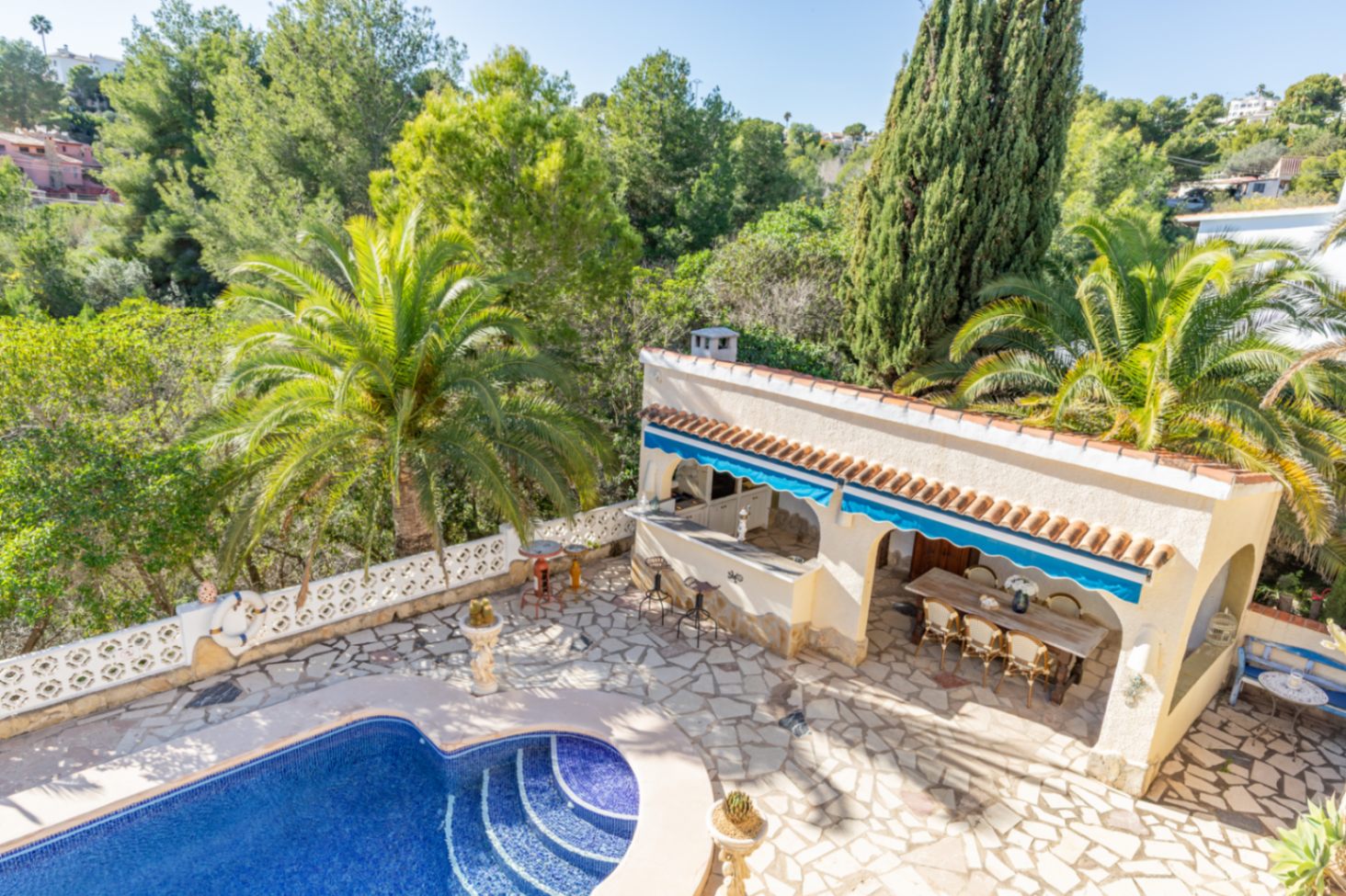 Espace extérieur comprenant une piscine en mosaïque, une terrasse en pierre et une cuisine d'été couverte, le tout niché dans un jardin méditerranéen arboré.