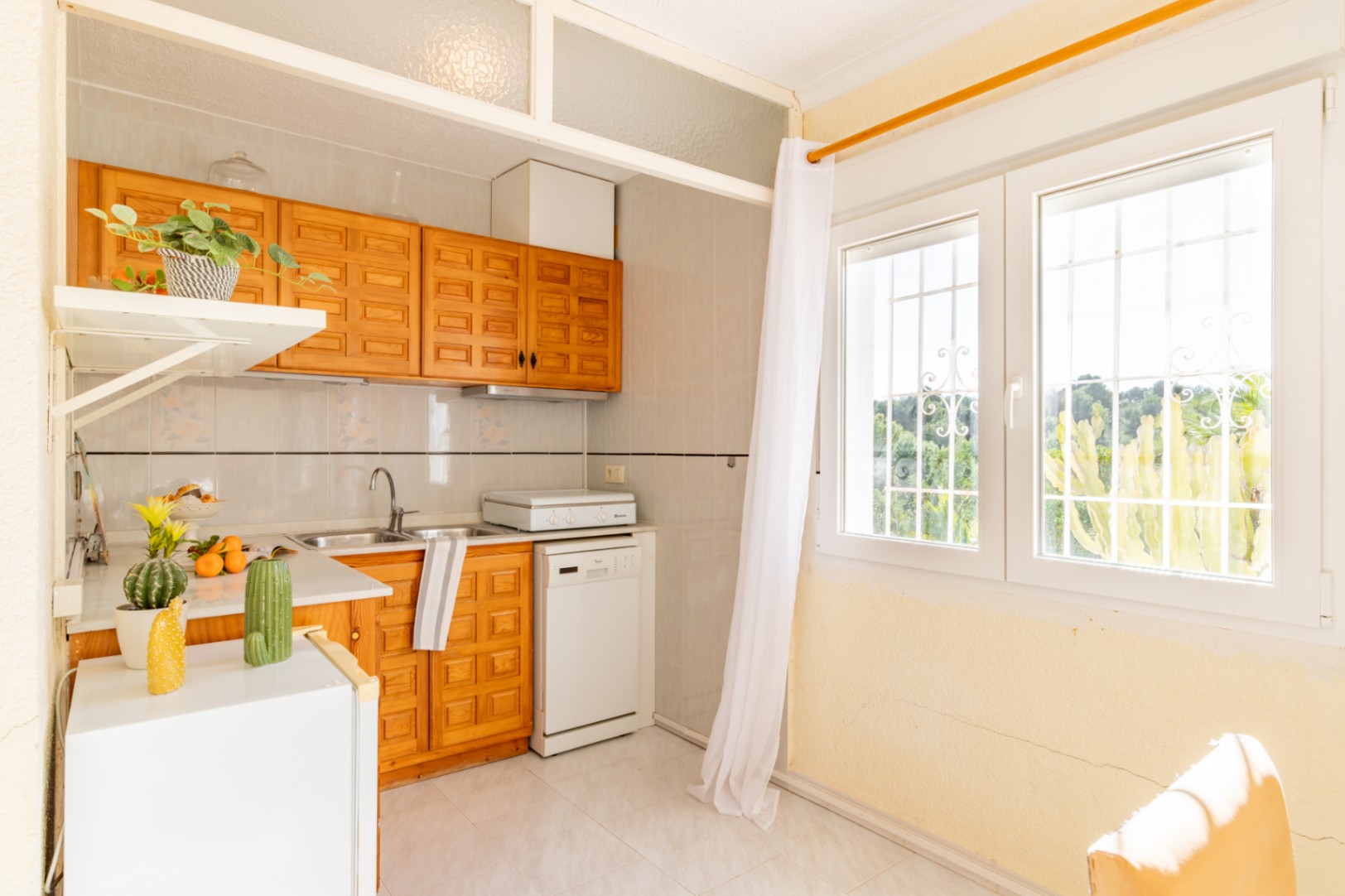 Kitchenette fonctionnelle de l'appartement indépendant avec placards en bois, plan de travail blanc et grande fenêtre offrant une lumière naturelle.