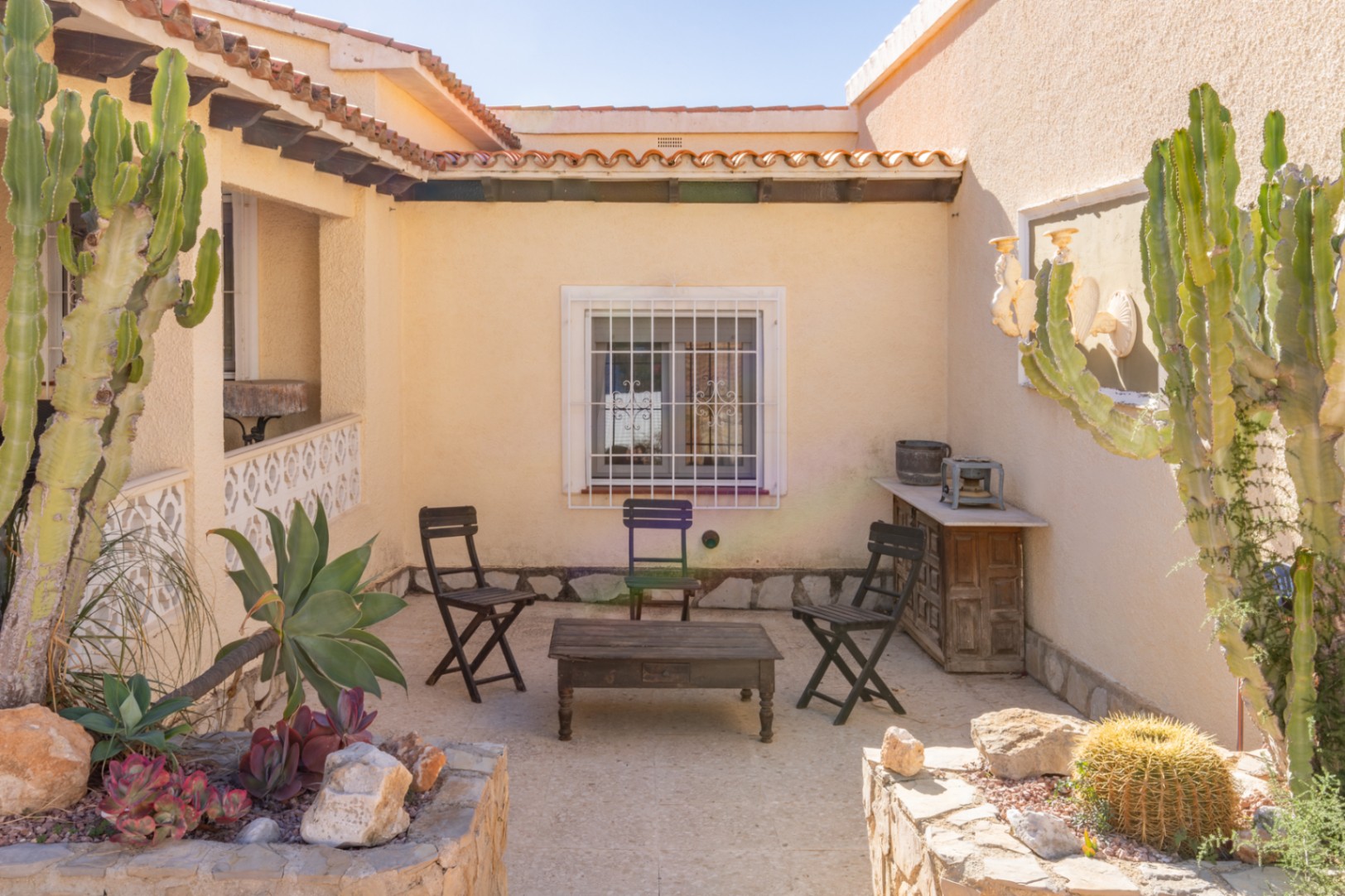 Patio extérieur de style méditerranéen avec sol en pierre, mobilier en bois rustique et jardinières intégrées plantées de cactus et de succulentes.