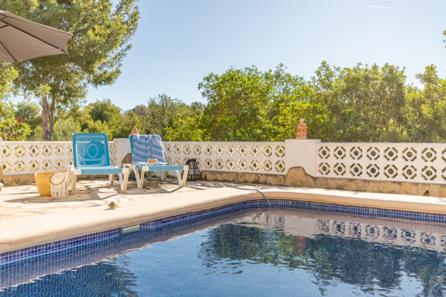Piscine en mosaïque bleue avec terrasse en pierre naturelle, bordée d'un mur de clôture ajouré blanc et d'une végétation méditerranéenne dense.