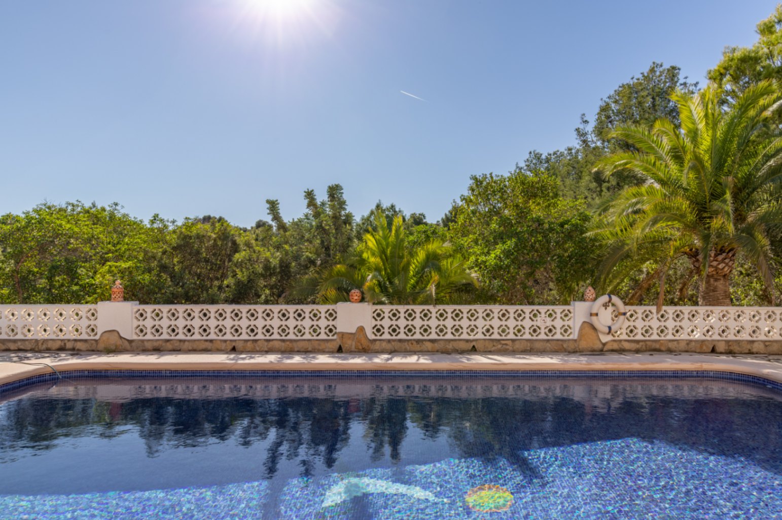 Piscine en mosaïque bleue orientée plein sud, bordée d'une balustrade blanche décorative et entourée d'une végétation méditerranéenne luxuriante.