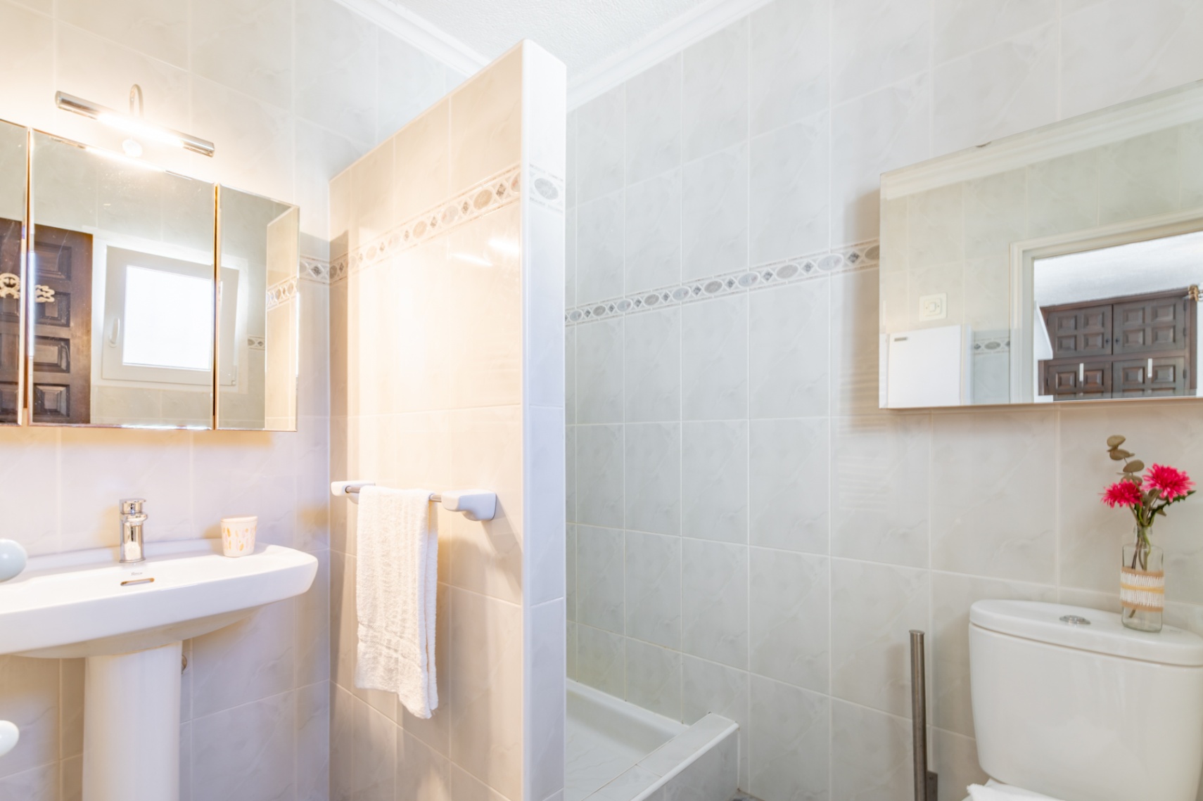 Salle de bains avec carrelage beige, lavabo sur colonne, armoire de toilette éclairée et douche séparée par une paroi fixe carrelée.