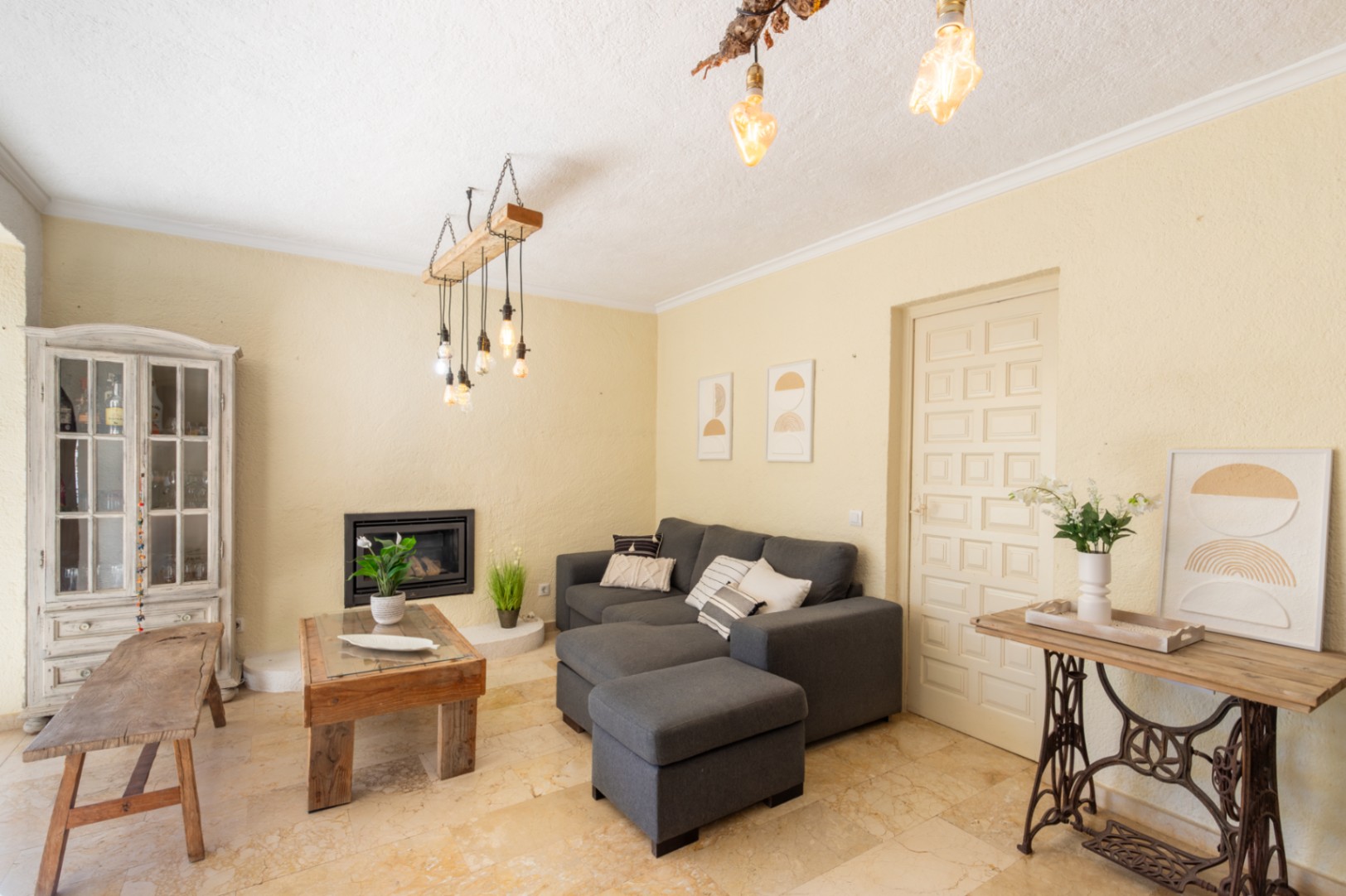 Salon avec cheminée encastrée, sol en marbre et mobilier rustique. L'espace se distingue par ses luminaires suspendus originaux et sa console sur pied de machine à coudre ancienne.