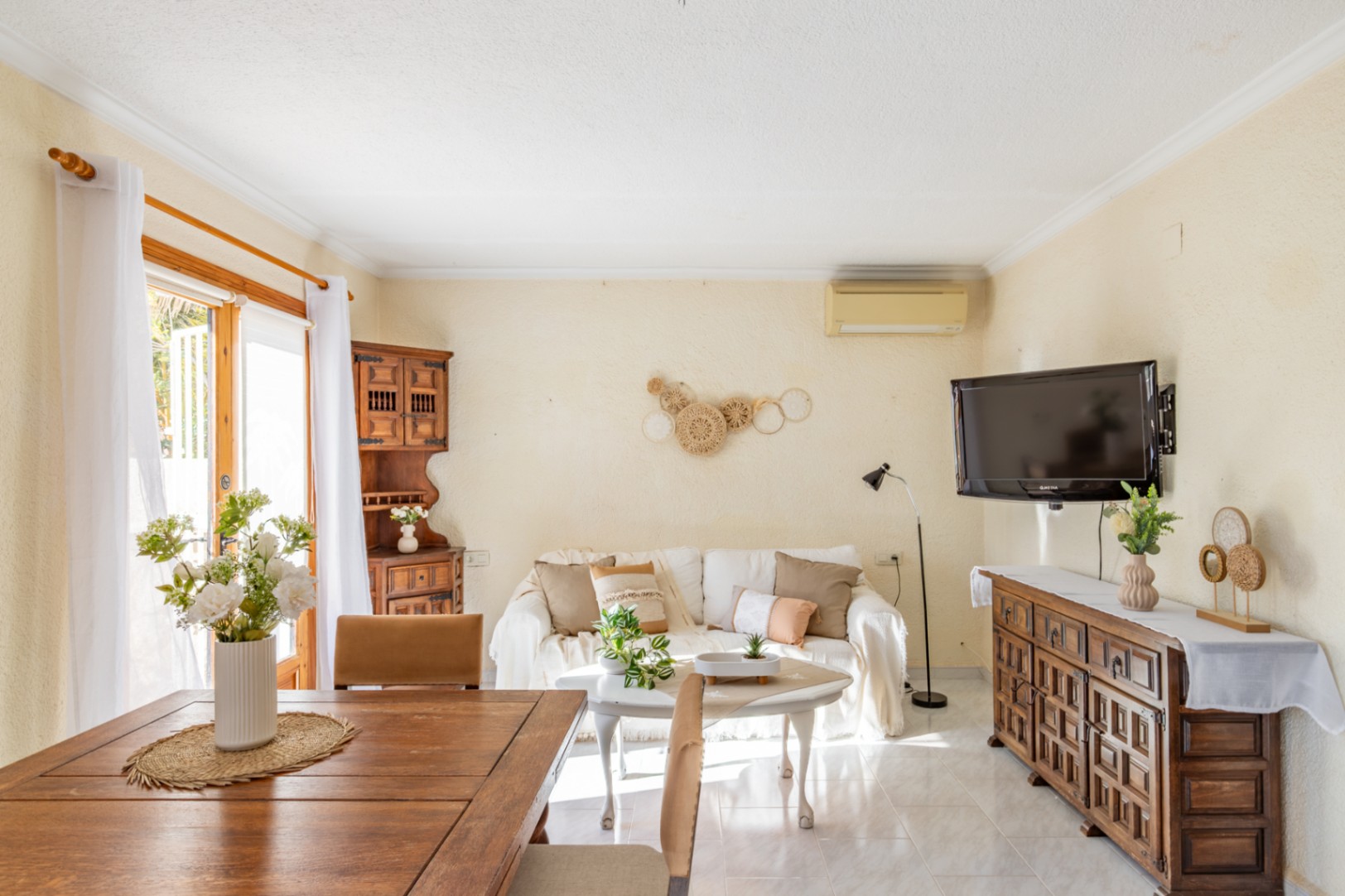 Salon-salle à manger lumineux avec carrelage clair, meubles en bois rustique et porte vitrée offrant une lumière naturelle abondante.