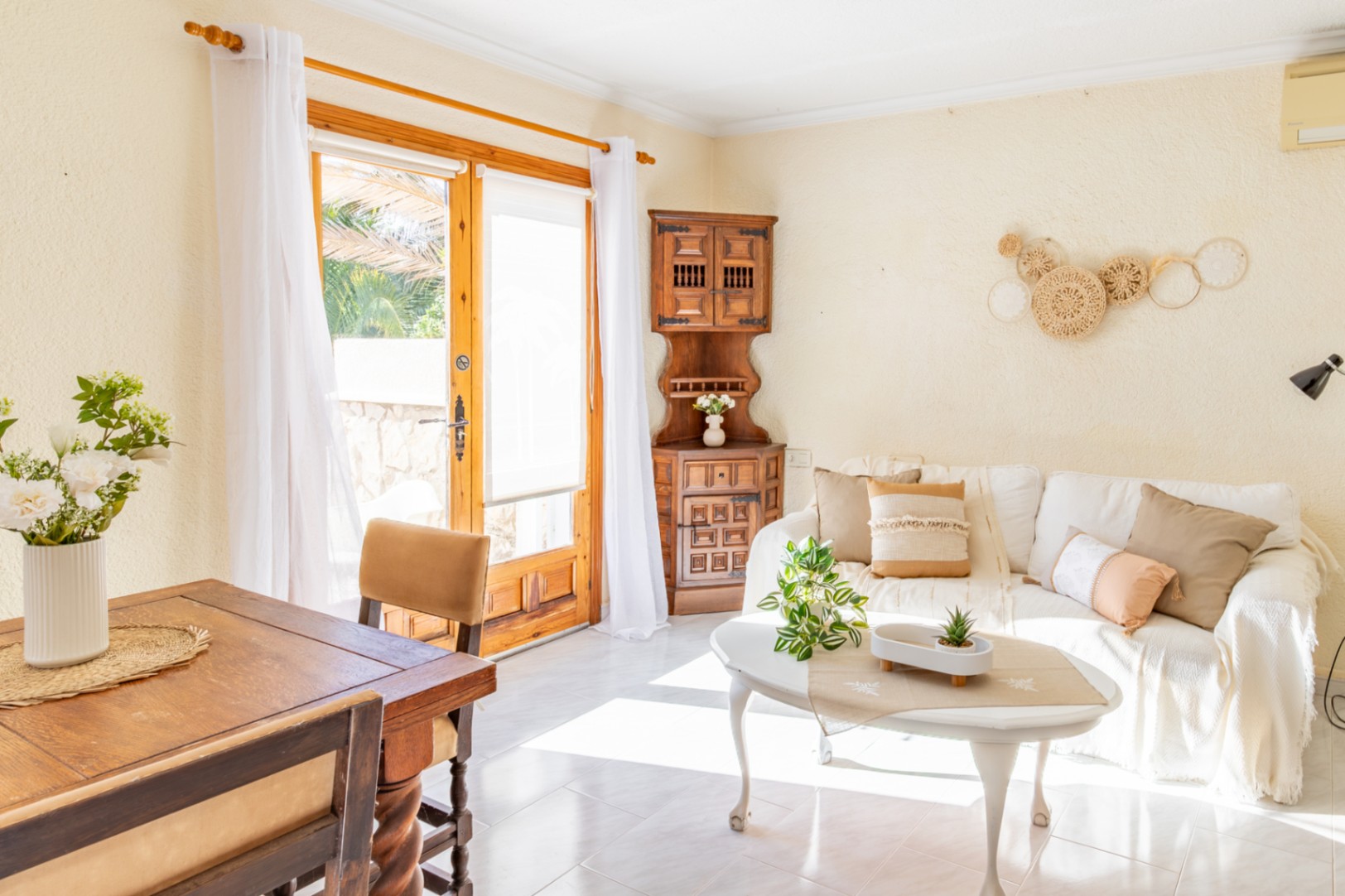 Salon-salle à manger lumineux avec mobilier en bois rustique, canapé blanc et accès direct à l'extérieur par une grande porte vitrée à cadre en bois naturel.