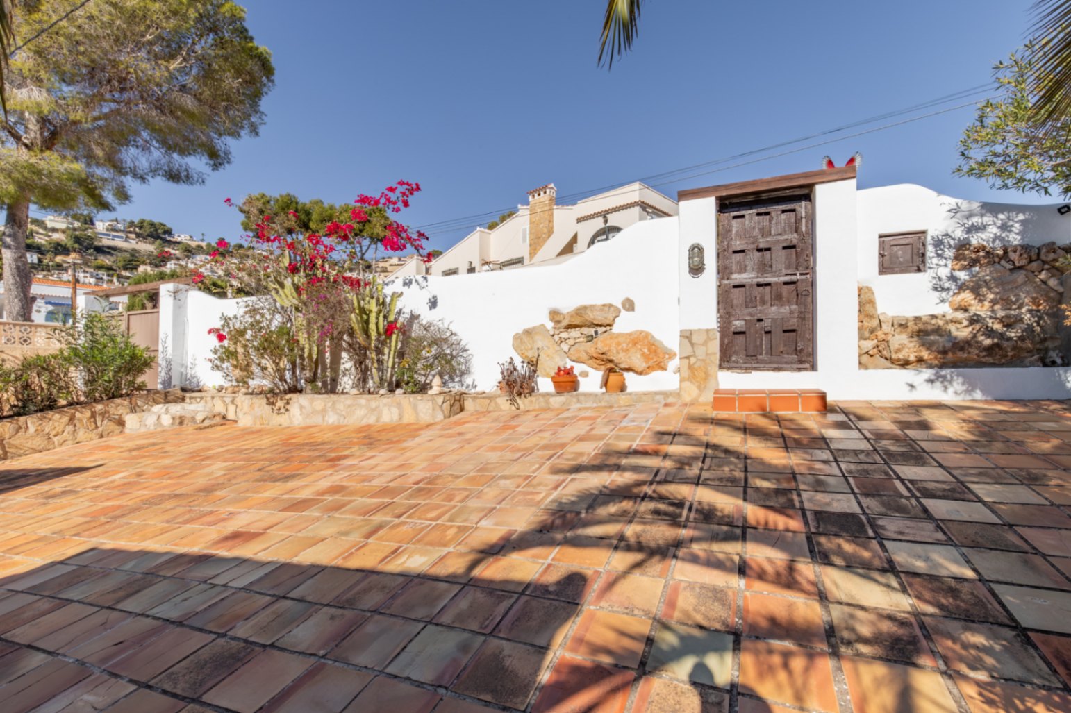 Terrasse extérieure avec sol en terre cuite, murs blancs ornés de pierres naturelles et un portail en bois rustique entouré de bougainvilliers et de cactus.