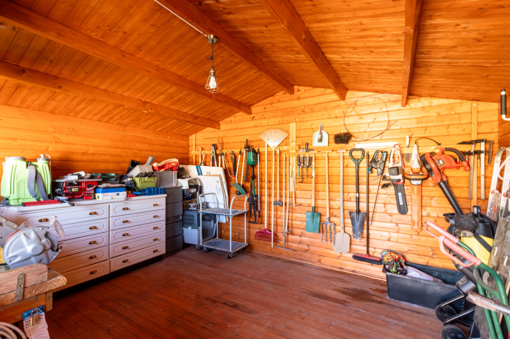 Abri de jardin en bois organisé, comprenant un rangement mural pour outils, une commode blanche et une structure à poutres apparentes.