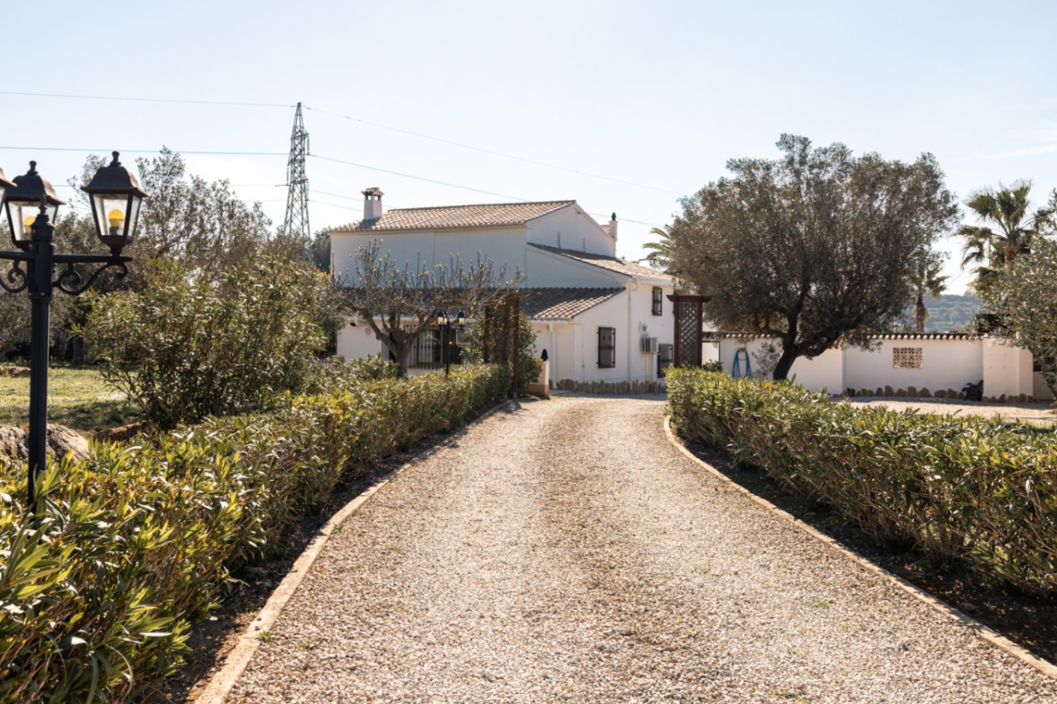 Allée de gravier bordée de haies basses menant à la façade blanche de la finca, illustrant l'architecture méditerranéenne traditionnelle et son jardin arboré.