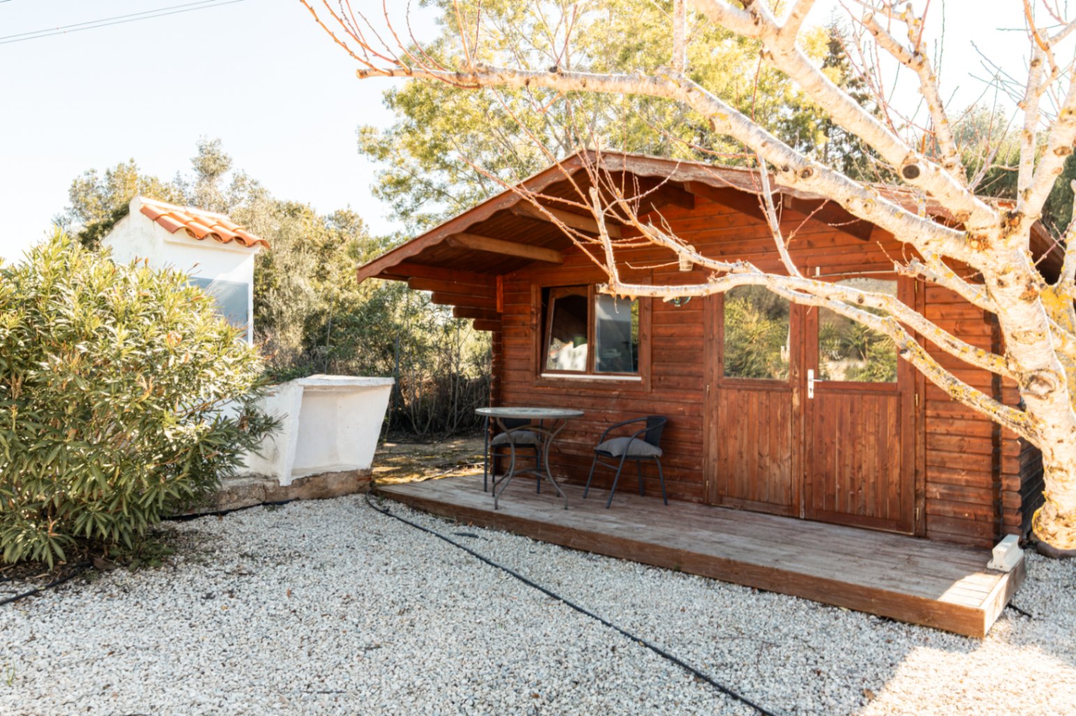 Cabane de jardin en bois avec terrasse surélevée, portes vitrées et coin salon, située dans un espace paysager avec gravier et arbres.