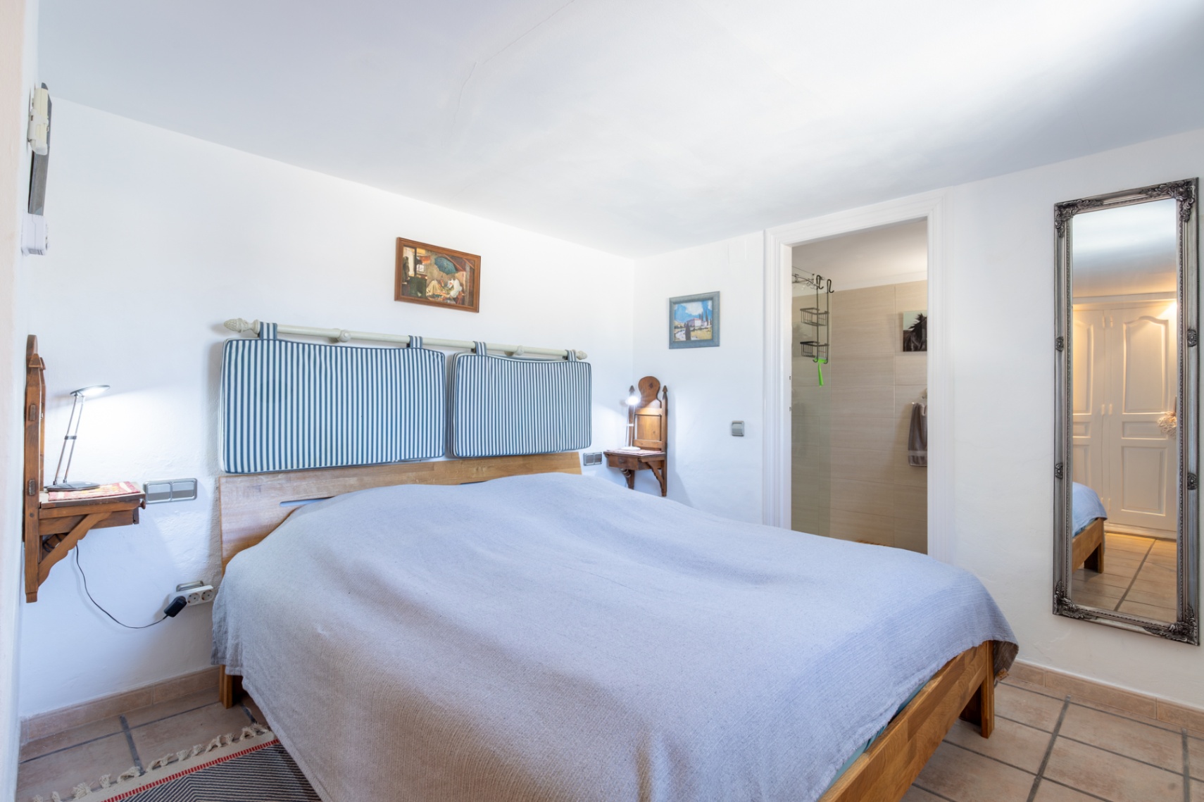 Chambre avec lit en bois, coussins de tête de lit rayés et tables de chevet murales. Elle dispose d'un accès direct à la salle de bain attenante et d'un grand miroir ornemental.