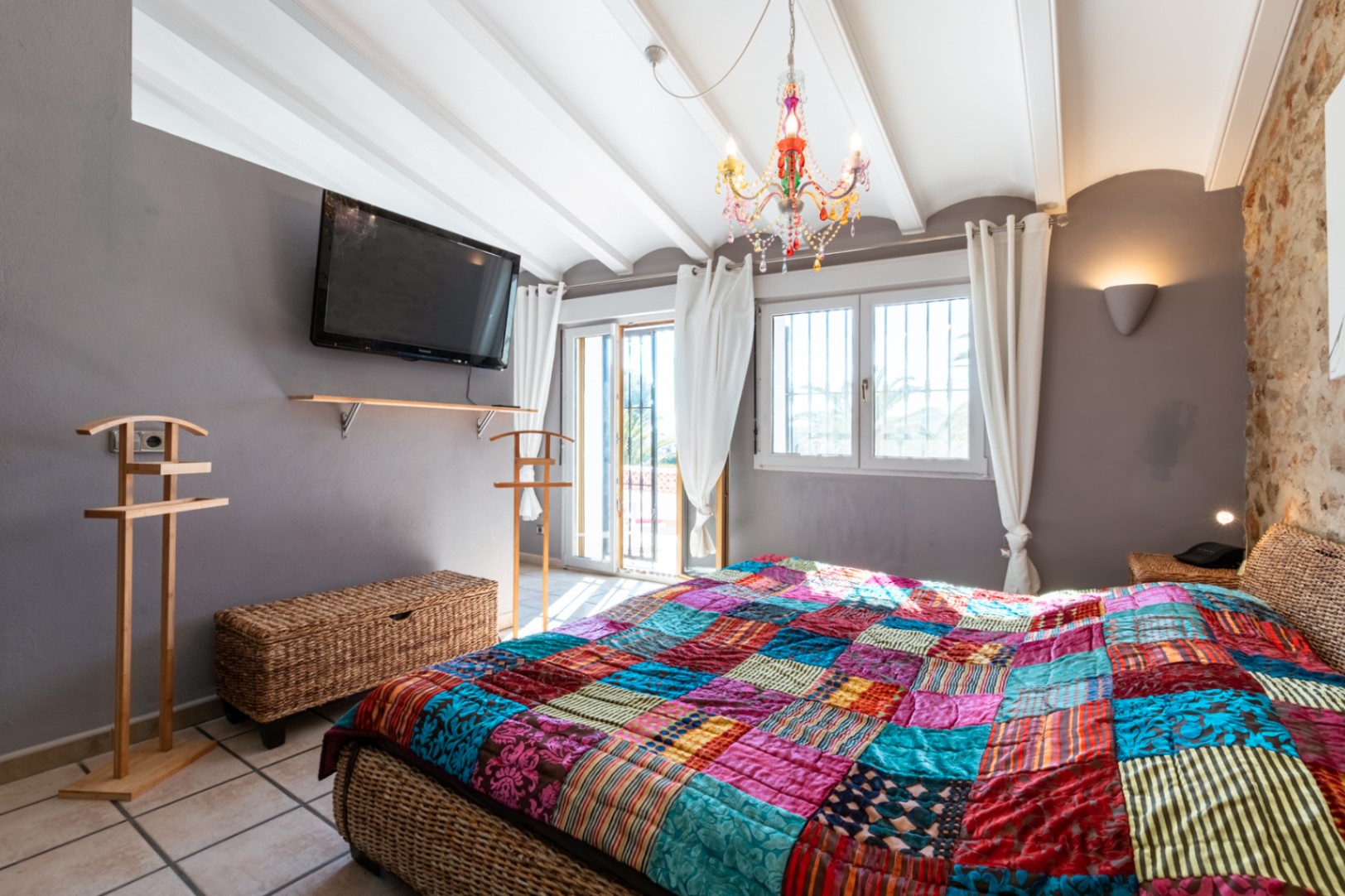 Chambre avec mur en pierre naturelle, plafond à poutres blanches et lit en osier. Elle dispose d'un lustre coloré et d'un accès direct vers l'extérieur.