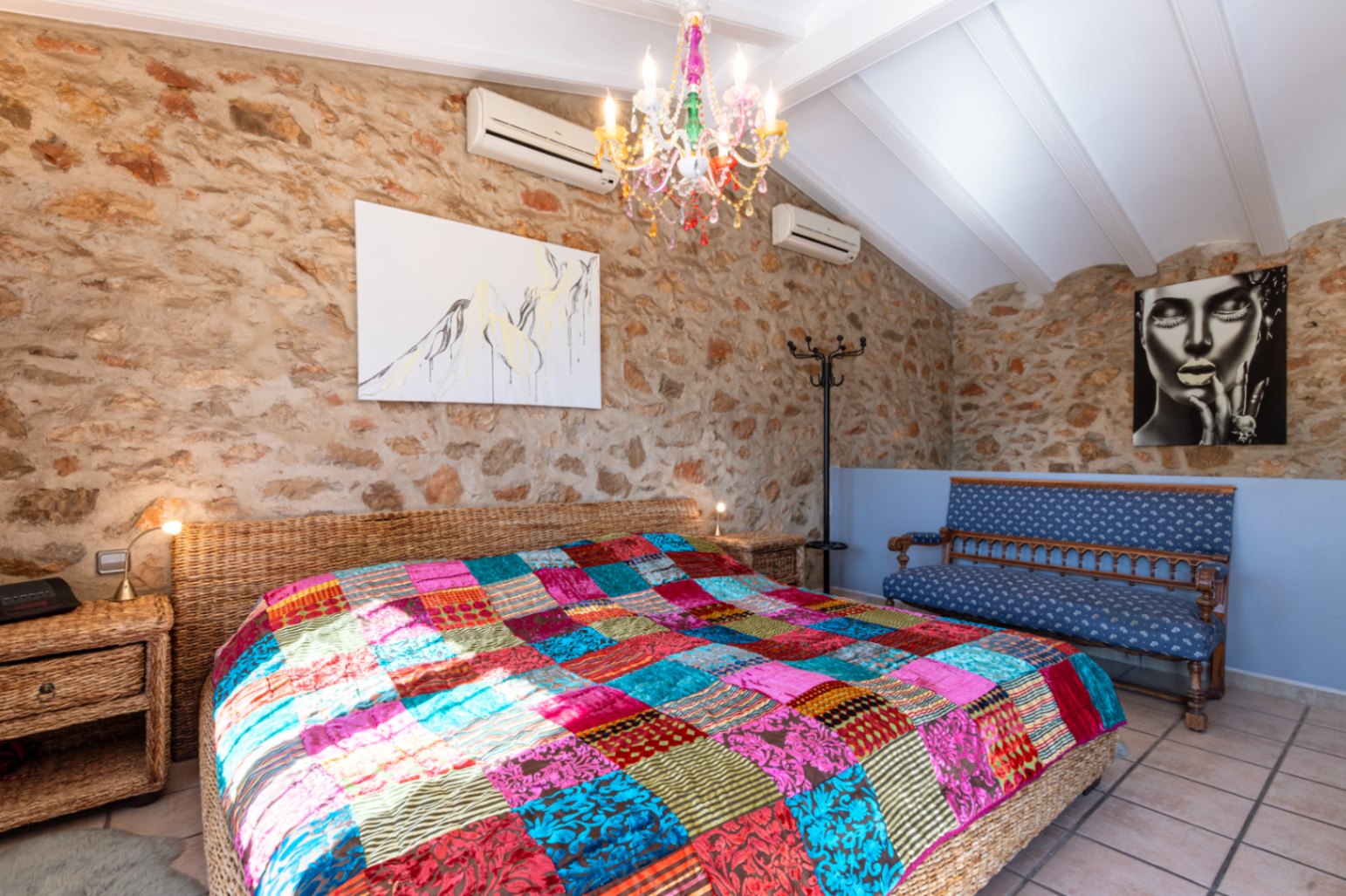 Chambre avec murs en pierre naturelle, plafond incliné à poutres blanches et lit en osier orné d'un couvre-lit en patchwork coloré sous un lustre en cristal.