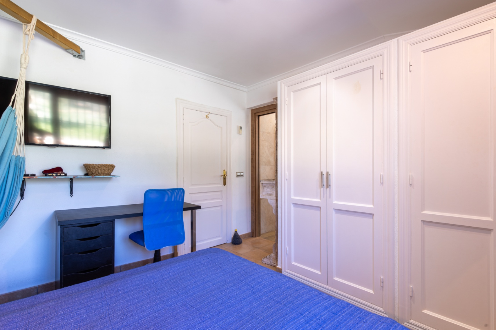 Chambre avec placards intégrés blancs, coin bureau avec chaise bleue et accès à une salle de bain attenante aux finitions en pierre.