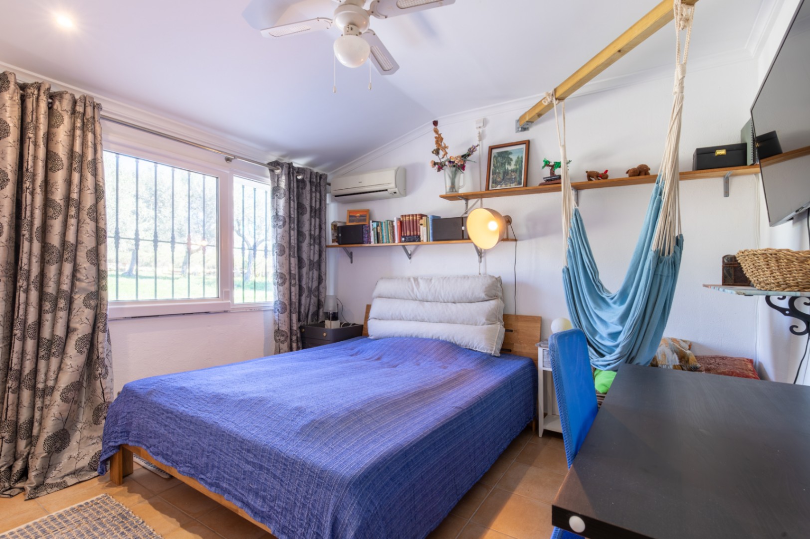 Chambre fonctionnelle avec lit double, coin bureau, étagères en bois et hamac suspendu. Comprend la climatisation, un ventilateur de plafond et une belle luminosité.